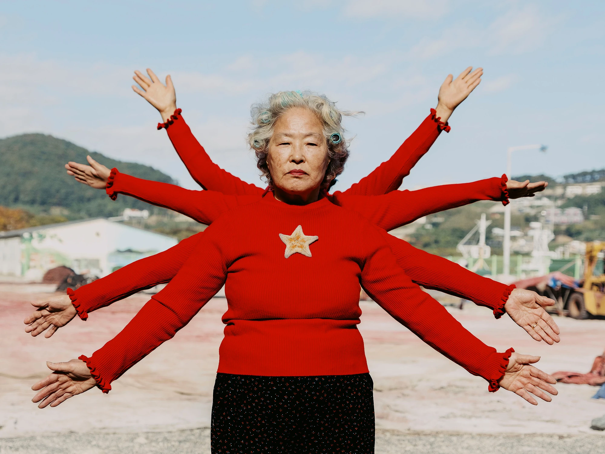 Nirvana by Daesung Lee, photographed by Daesung Lee — A woman in a star-emblazoned red sweater appears with six arms fanned wide, a many-armed deity standing in a bare industrial yard.
