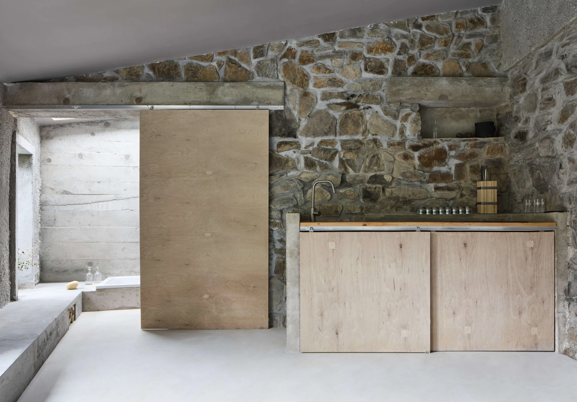 Panoši Újezd Summer House by Lenka Milerová, photographed by Radek Šrettr Úlehla — A plywood sliding door and run of low plywood kitchen cabinets stand against a tall rough stone wall beside board-formed concrete.