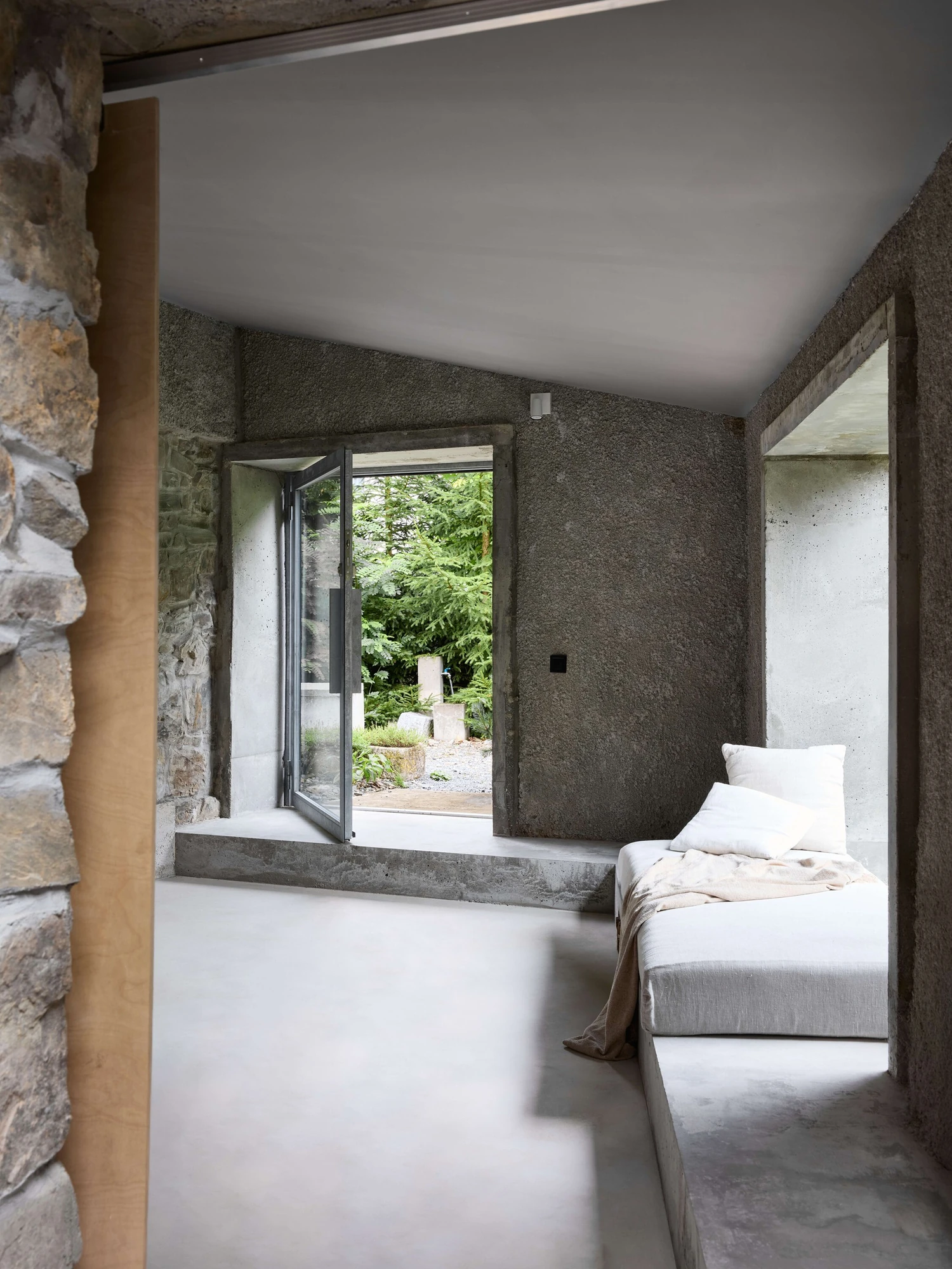 Panoši Újezd Summer House by Lenka Milerová, photographed by Radek Šrettr Úlehla — A linen-topped concrete daybed runs beneath an open pivot window, a plywood doorway at left and rough stone framing the view.