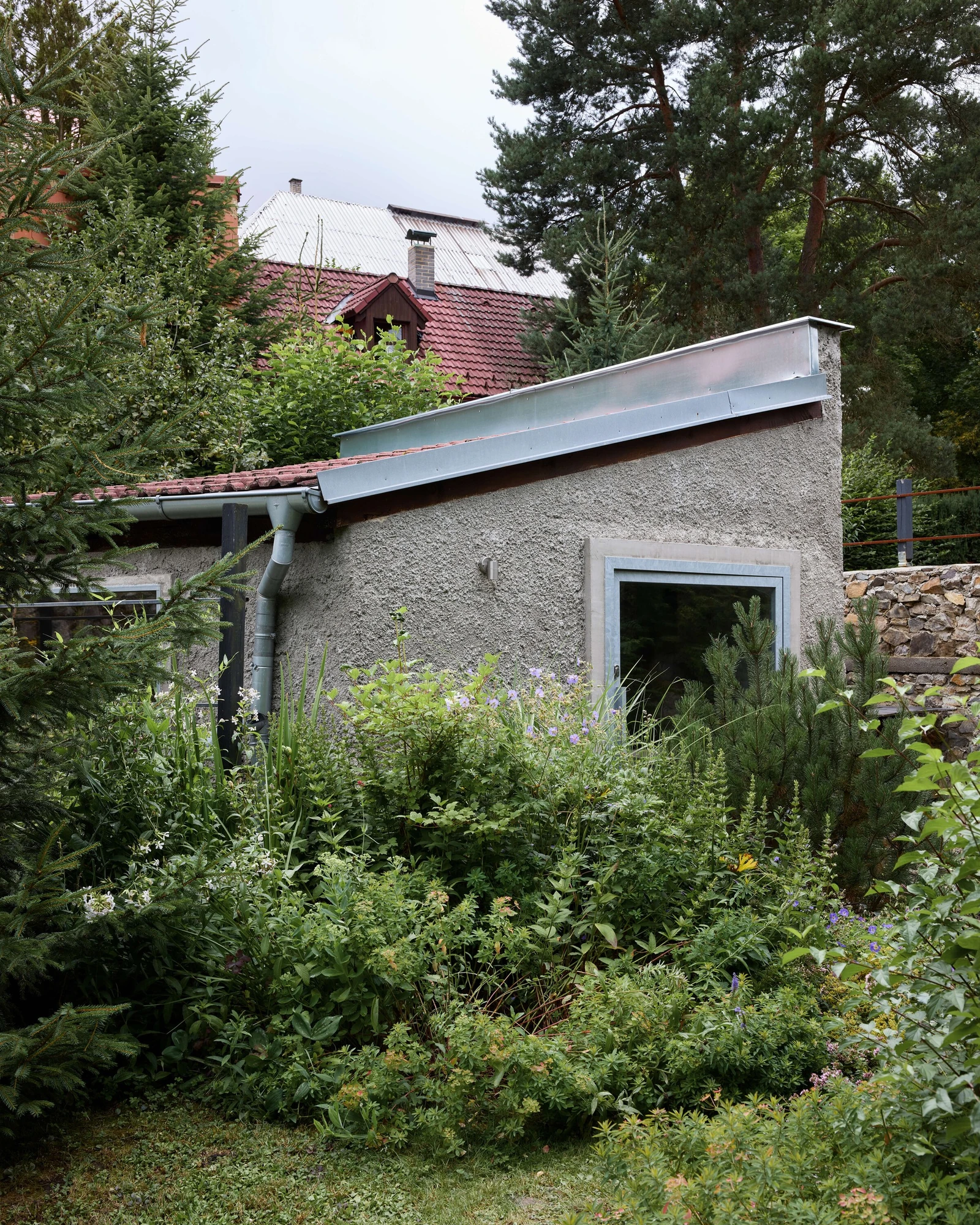 Panoši Újezd Summer House by Lenka Milerová, photographed by Radek Šrettr Úlehla — The rendered grey gable rises from an overgrown garden, a concrete-framed window and a folded sheet-metal roof edge marking the new work.