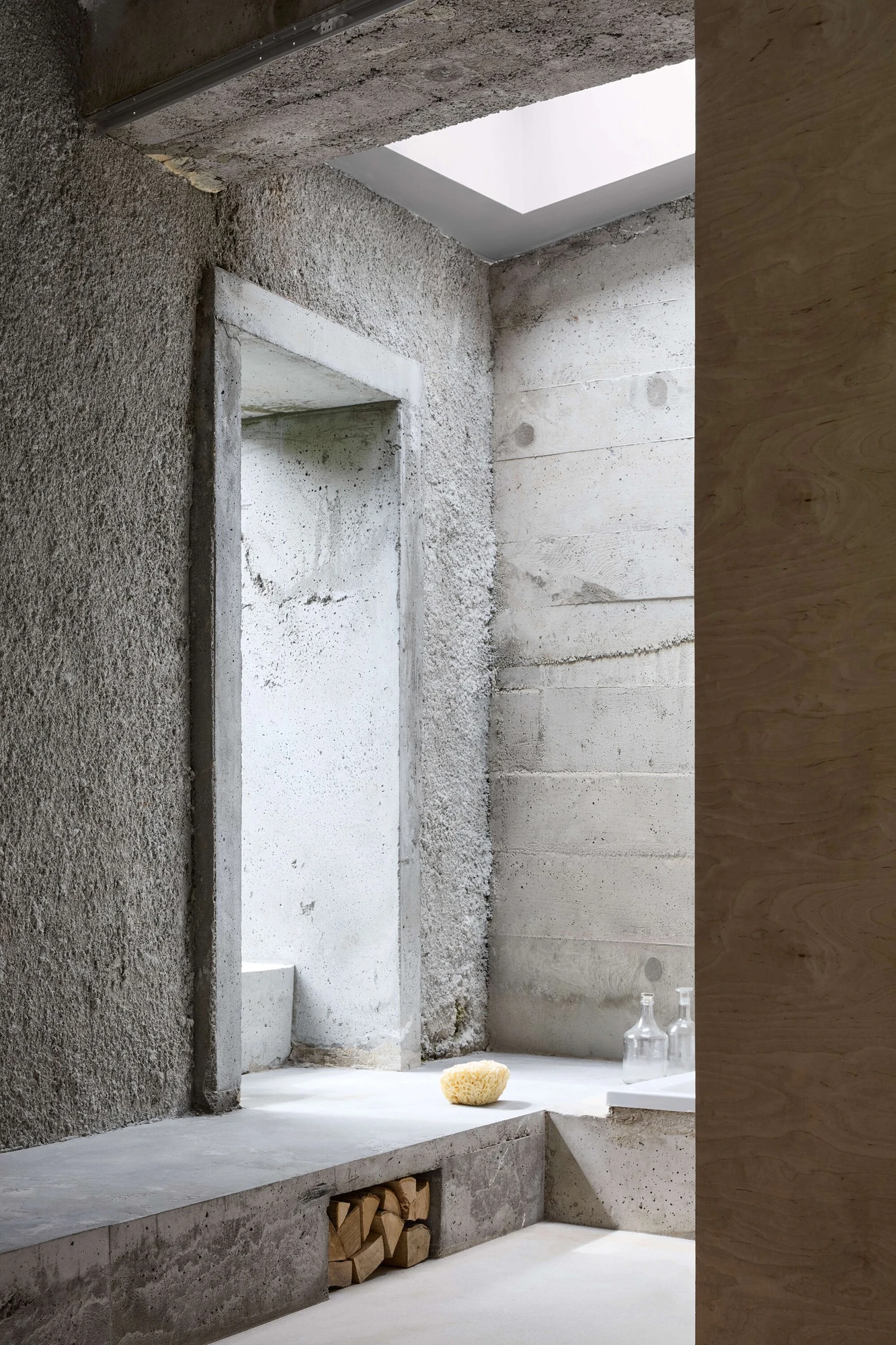 Panoši Újezd Summer House by Lenka Milerová, photographed by Radek Šrettr Úlehla — A tall recessed niche cut between rough render and board-formed concrete rises above the bench, a sponge and firewood resting below.