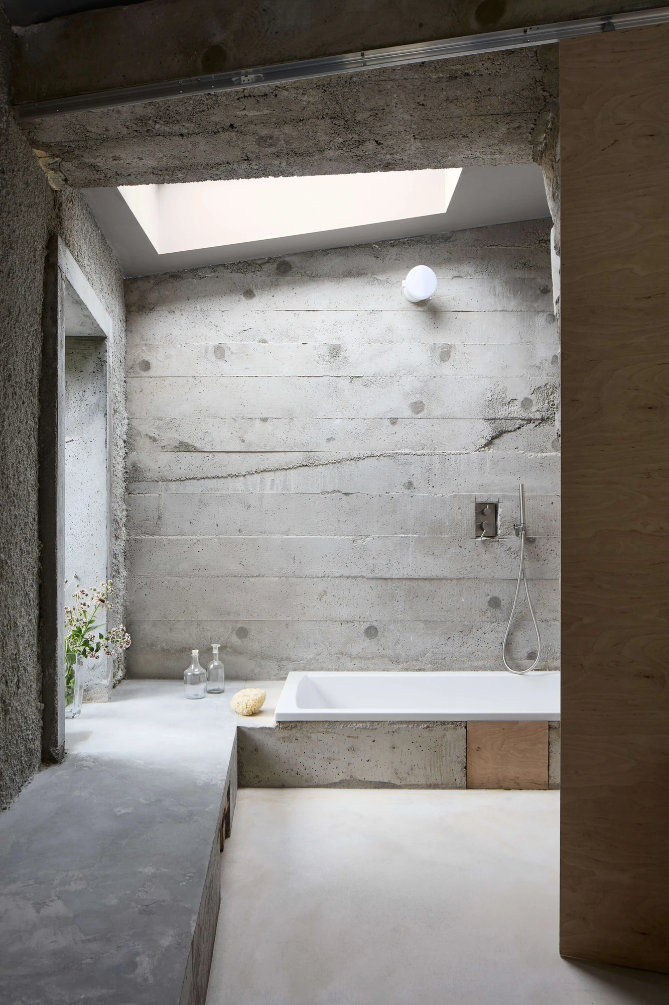 Panoši Újezd Summer House by Lenka Milerová, photographed by Radek Šrettr Úlehla — A bathtub set into a concrete surround sits below a skylight, a board-formed concrete wall behind and a plywood panel closing the side.