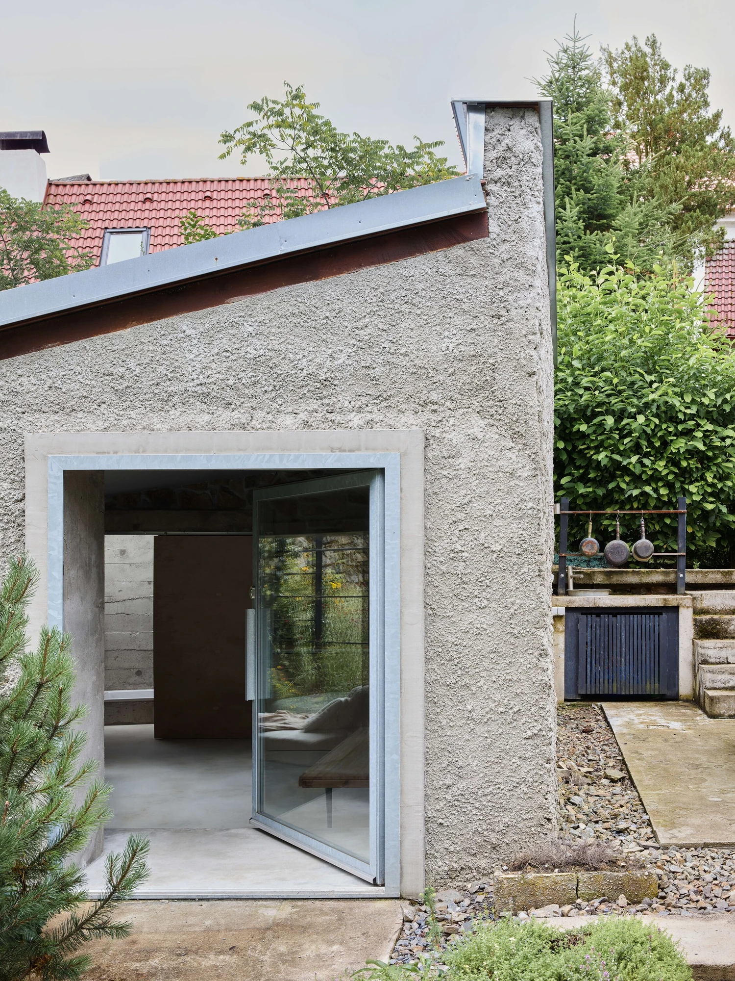 Panoši Újezd Summer House by Lenka Milerová, photographed by Radek Šrettr Úlehla — A glazed pivot door framed in a thick concrete surround opens at the building's corner beneath the pitched render wall.
