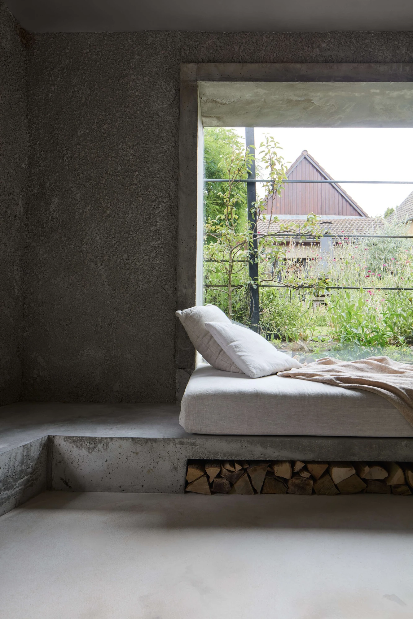 Panoši Újezd Summer House by Lenka Milerová, photographed by Radek Šrettr Úlehla — A concrete window seat with a linen mattress and split firewood stacked beneath faces the garden through a steel-railed opening.