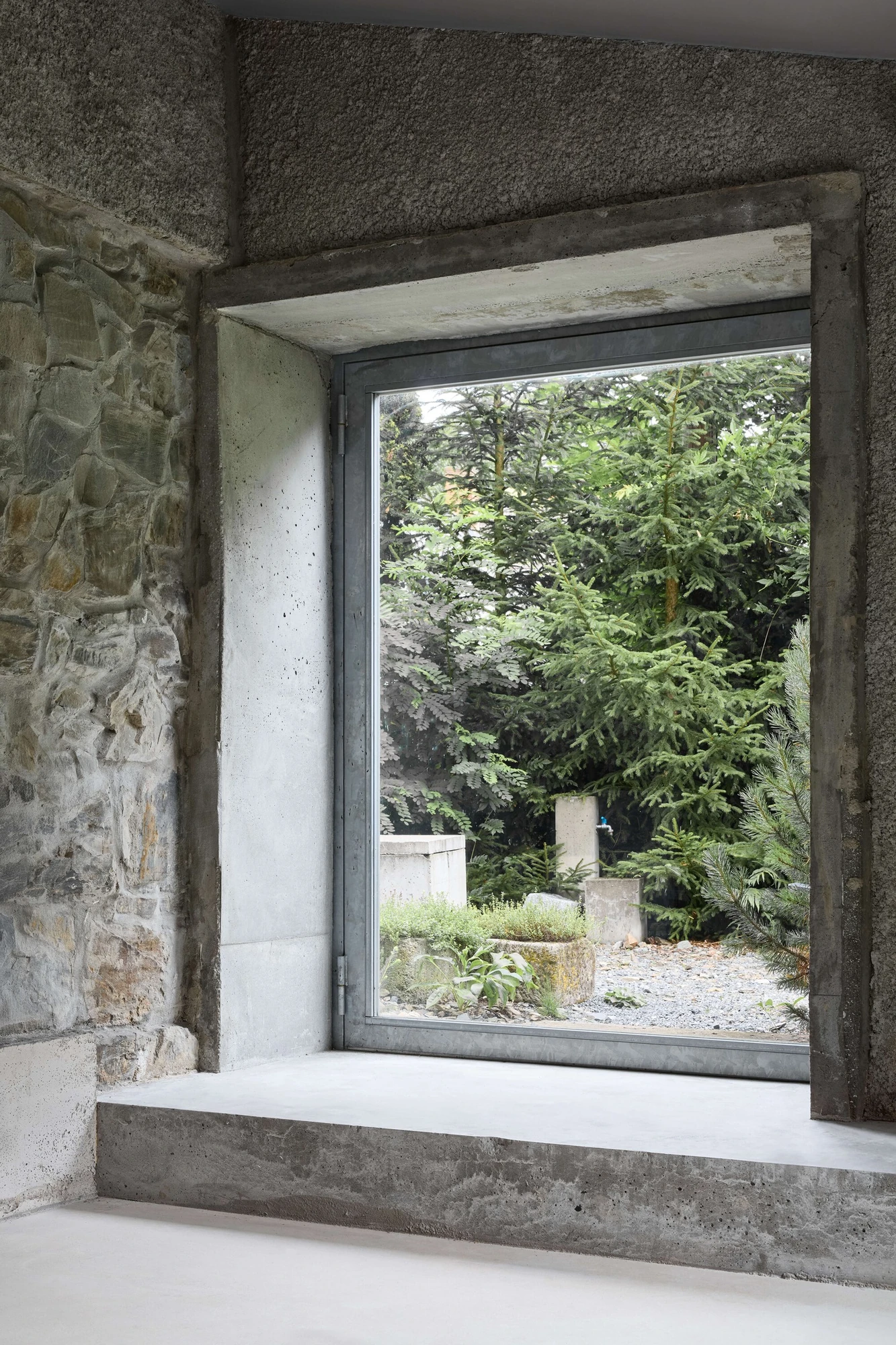 Panoši Újezd Summer House by Lenka Milerová, photographed by Radek Šrettr Úlehla — A deep concrete-lined window cut through rough stone masonry frames stone garden ornaments and conifers beyond.