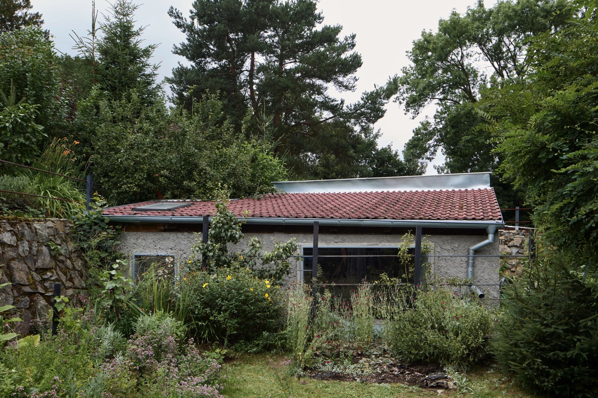 Panoši Újezd Summer House by Lenka Milerová, photographed by Radek Šrettr Úlehla — The full garden elevation, terracotta-tiled and pebbledash-rendered, sits low beneath pines with a wide glazed opening at its centre.
