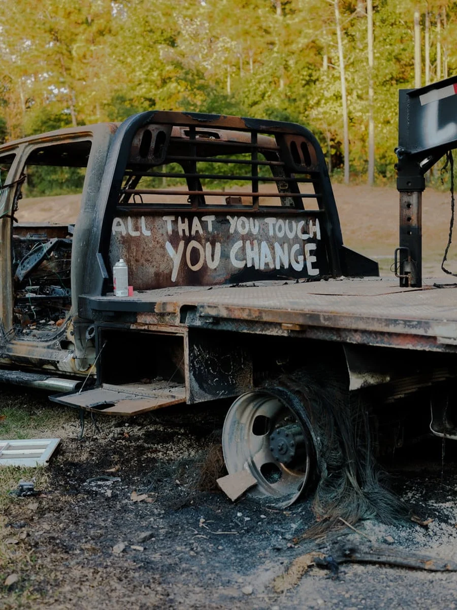 It's Not the Wind by Jordan Conway, photographed by Jordan Conway — A burnt-out flatbed truck stands in a clearing, ALL THAT YOU TOUCH YOU CHANGE painted in white across its charred bed.
