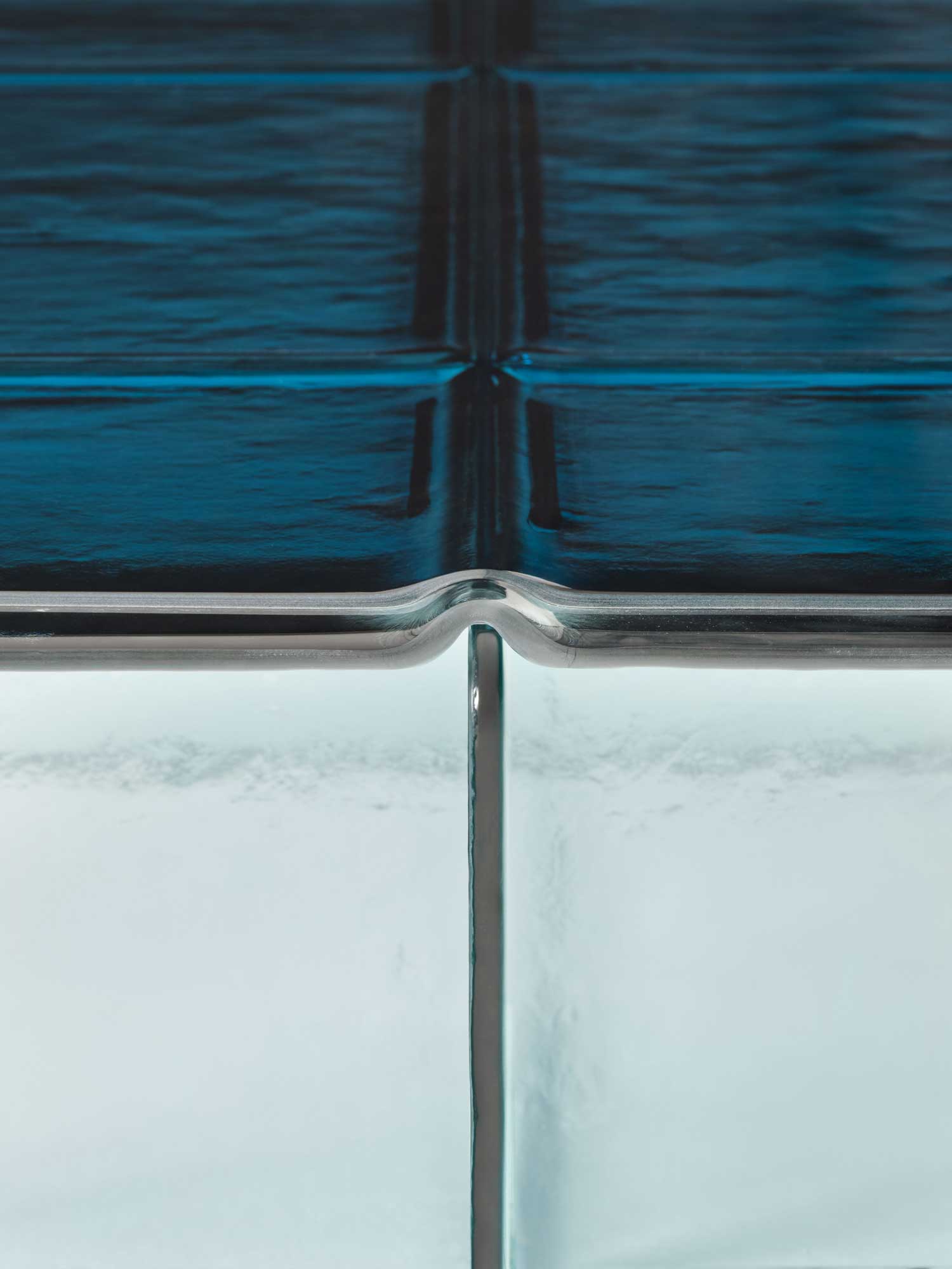 Glacier by Patricia Urquiola — Macro of a blue Glacier top where two cast tiles meet, the glossy surface reading almost black with wet, wave-like reflections above a teal glass fin.