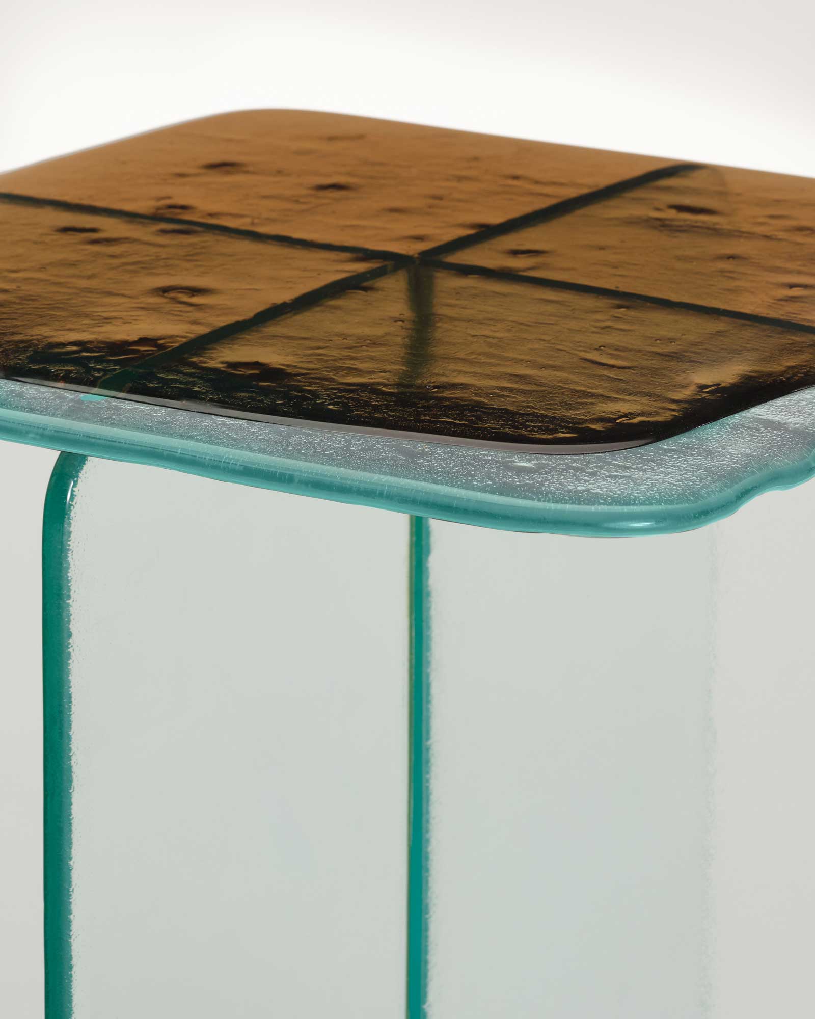 Glacier by Patricia Urquiola — Two Glacier side tables side by side, one charcoal-grey and one bronze-amber, their cast tops carried on crossed vertical fins of clear laminated glass.