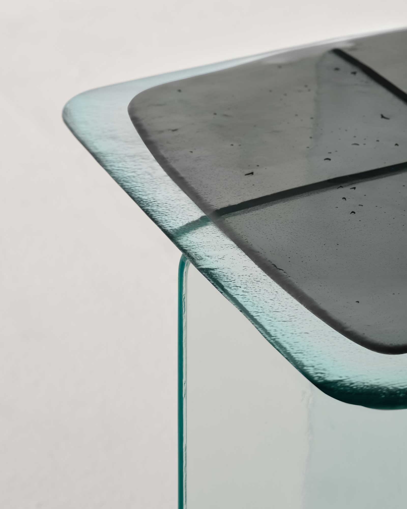 Glacier by Patricia Urquiola — Macro corner of a grey Glacier side table, the smoky cast top pitted with fine bubbles and cut by a faint tile grid above the transparent base edge.