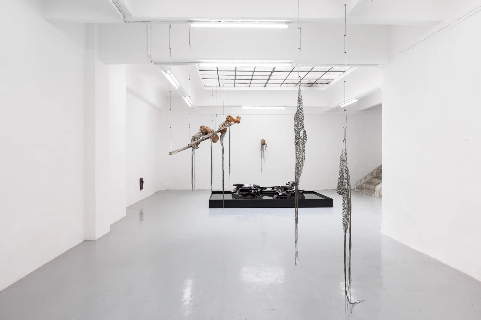 NADIR by Zishi Han and Laurence Sturla — Two beaded steel-wire nets hang the full height of a white gallery beside a length of London Plane wood, while a graphite-blackened cast sits in a shallow black pool on the polished floor.