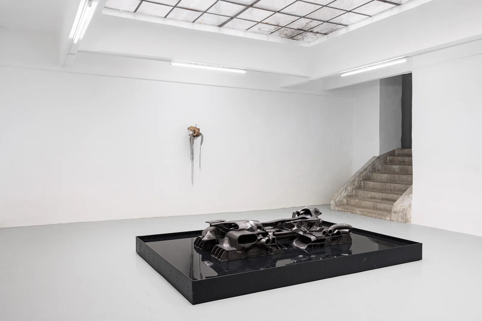 NADIR by Zishi Han and Laurence Sturla — A dark cast form, part engine and part terrain, sits in a black pool near a flight of raw concrete stairs.