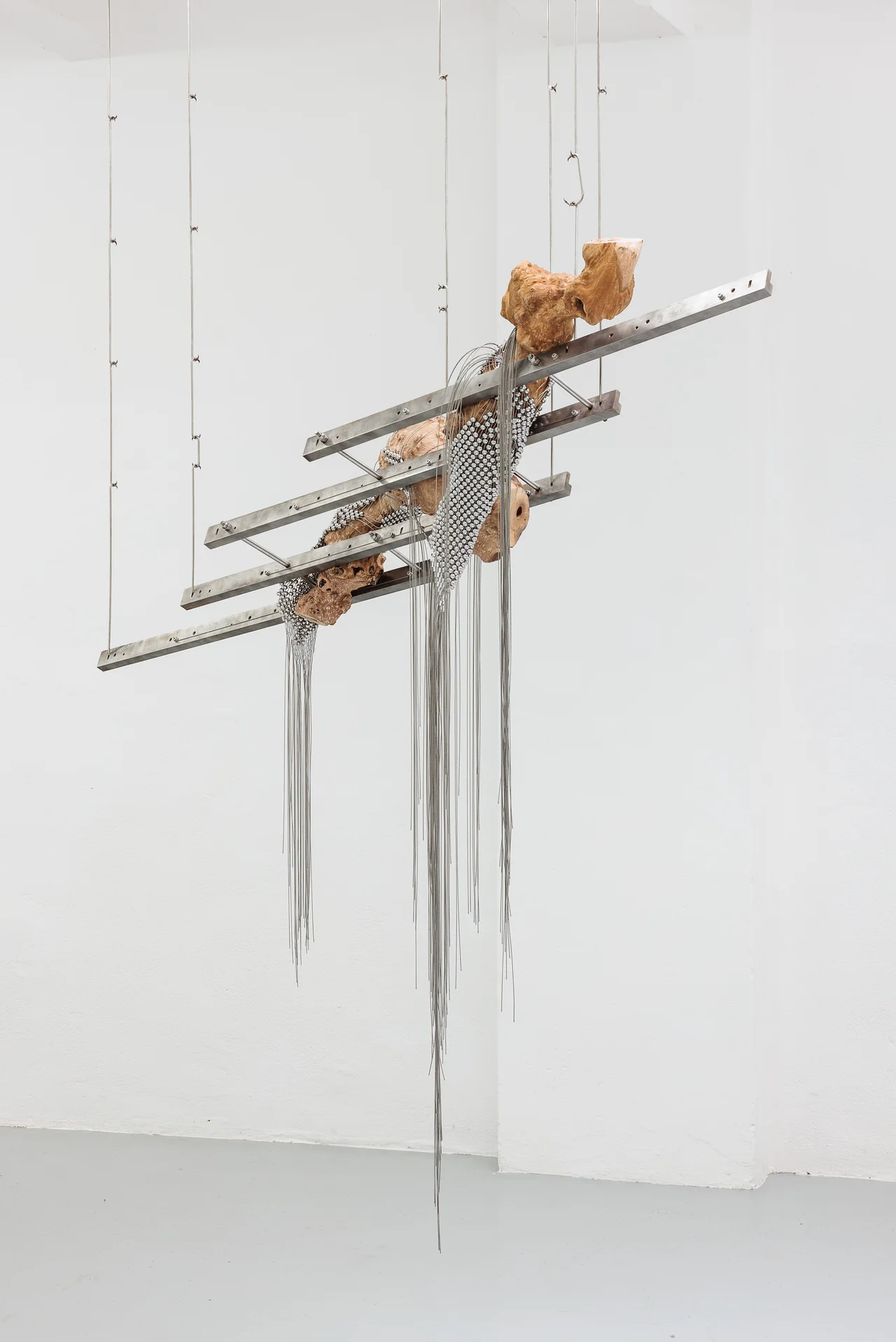 NADIR by Zishi Han and Laurence Sturla — Offcuts of London Plane wood are stacked across horizontal steel rails, draped in beaded netting that unravels into long silver wire below.
