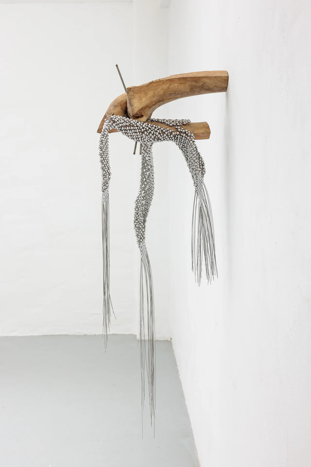 NADIR by Zishi Han and Laurence Sturla — A curved length of London Plane branch is bracketed to a white wall, a beaded steel-wire net slung over it with wire fringe hanging to the floor.