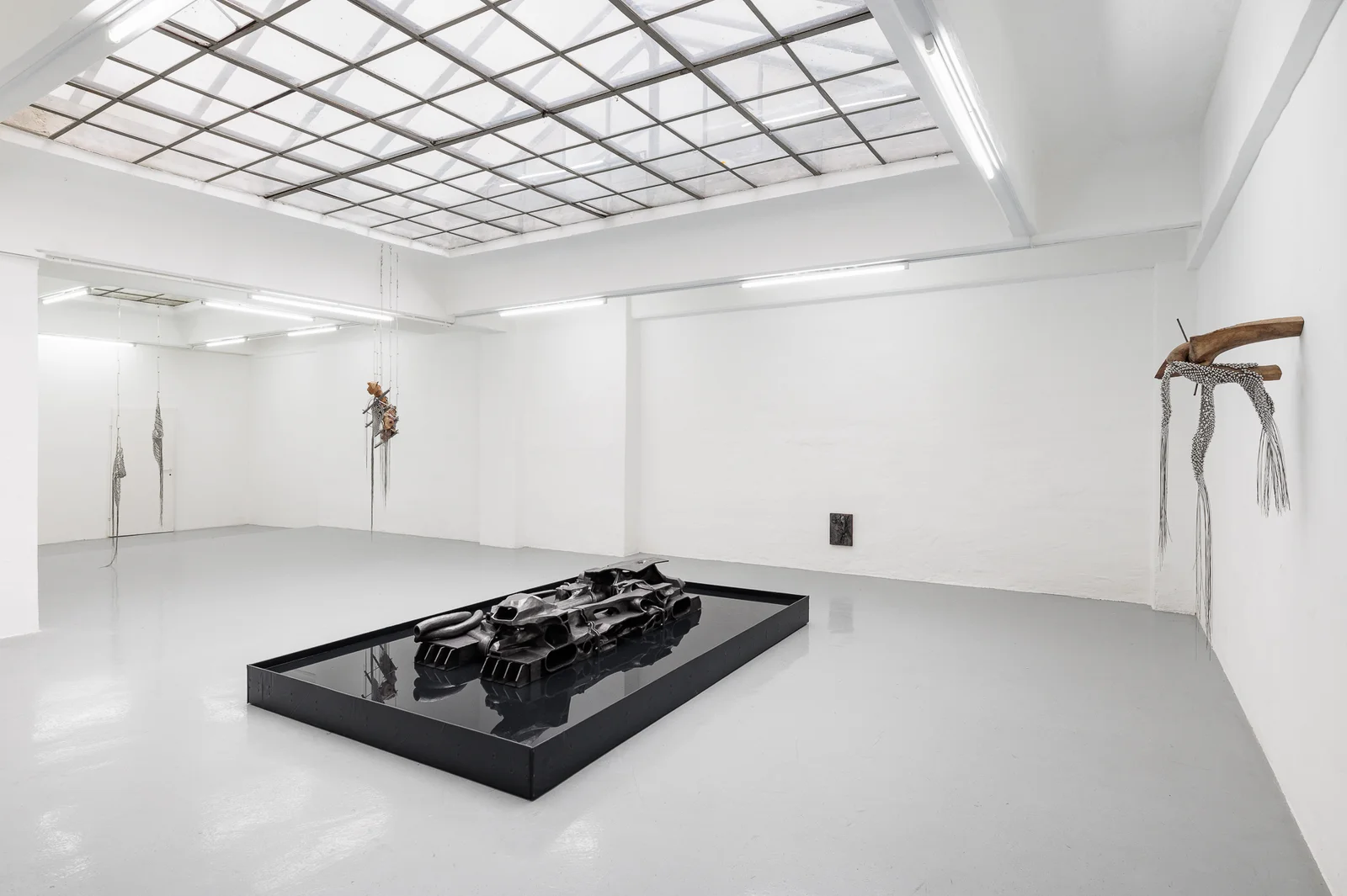 NADIR by Zishi Han and Laurence Sturla — Under a gridded skylight, a graphite-patinated ceramic form rests in a black reflecting pool at the centre of the gallery, hung sculptures at the far walls.