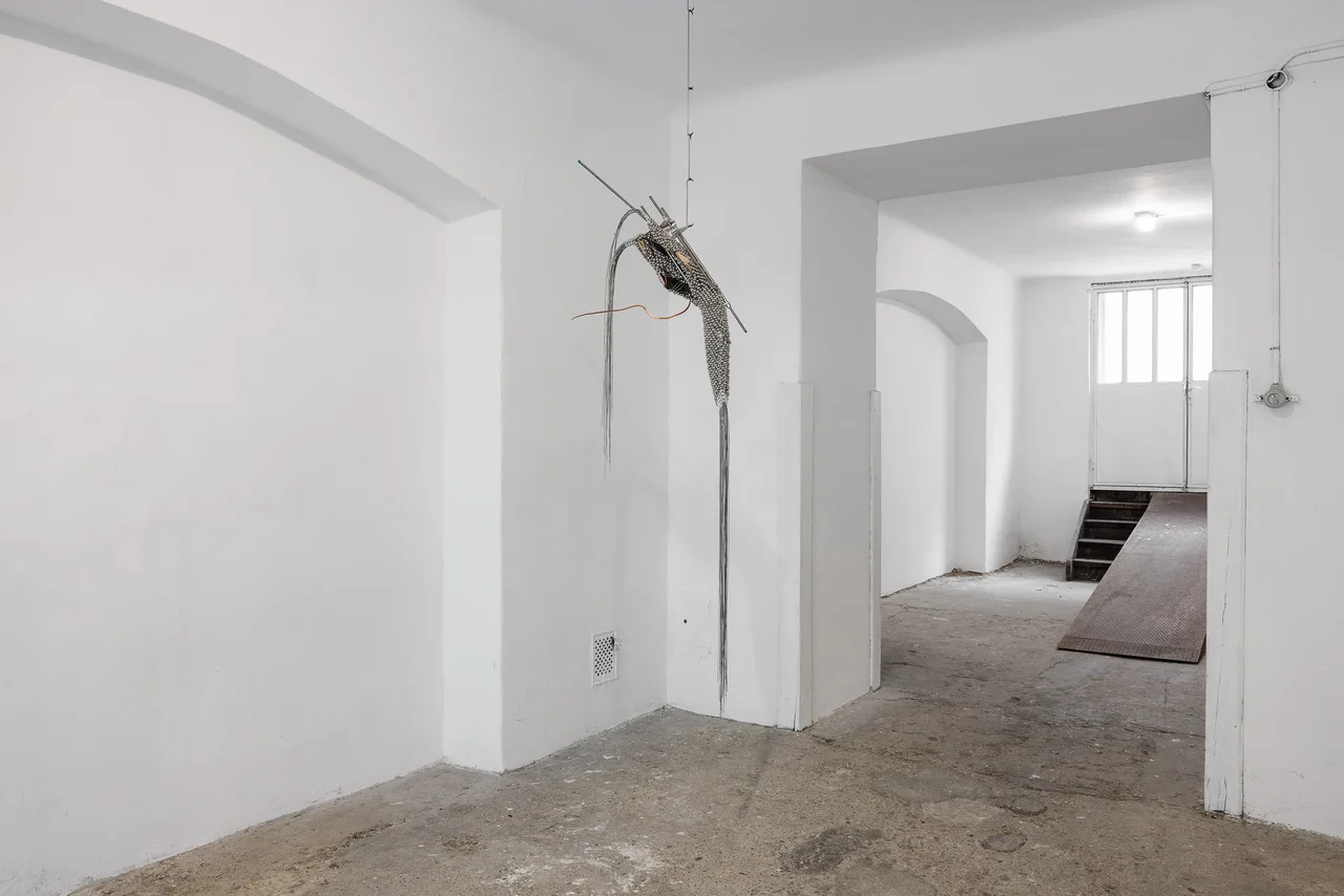 NADIR by Zishi Han and Laurence Sturla — In a white vaulted room with a raw concrete floor, a single beaded net and wood piece hangs near a doorway opening to daylight.