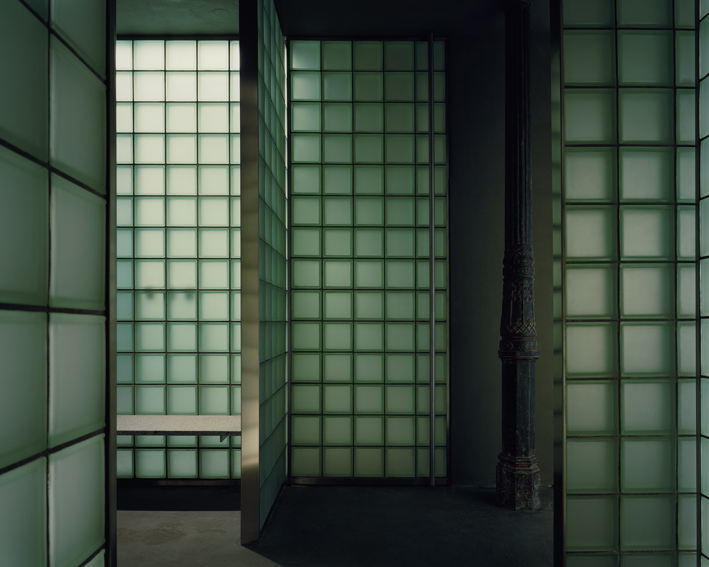 Riela by Oficina Satélite, photographed by Gregori Civera — Glass-block partitions frame an ornamented cast-iron column kept from the original building, a granite bench glowing behind the green cells, the floor dropping away into shadow.