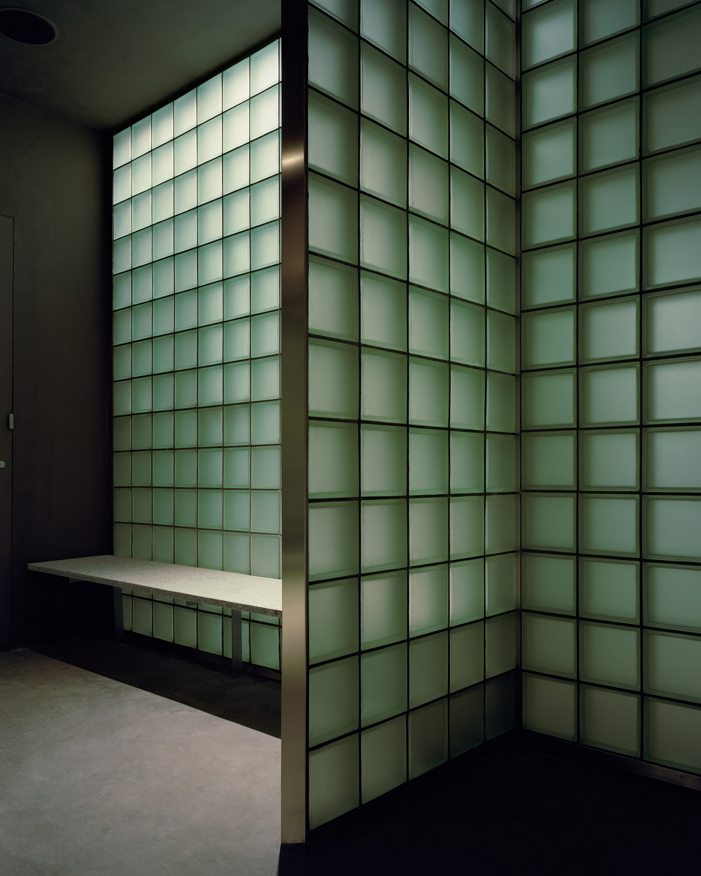 Riela by Oficina Satélite, photographed by Gregori Civera — A corner of pale-green glass block backlit from within, a slim granite bench cantilevered from the wall above a dark polished floor, a brass reveal marking the outer edge.