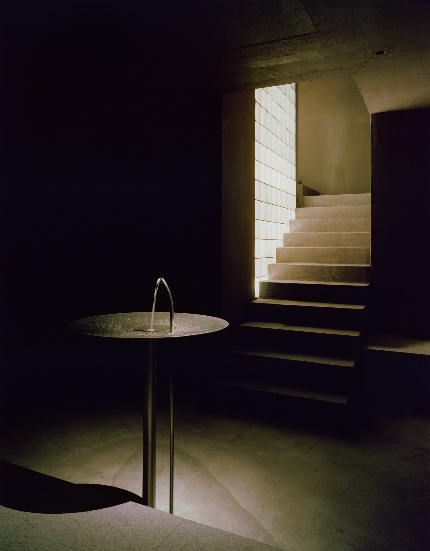 Riela by Oficina Satélite, photographed by Gregori Civera — The descent to water, pale concrete stairs lit by a glass-block wall leading down to a round granite fountain where a low arc of water falls back on itself.