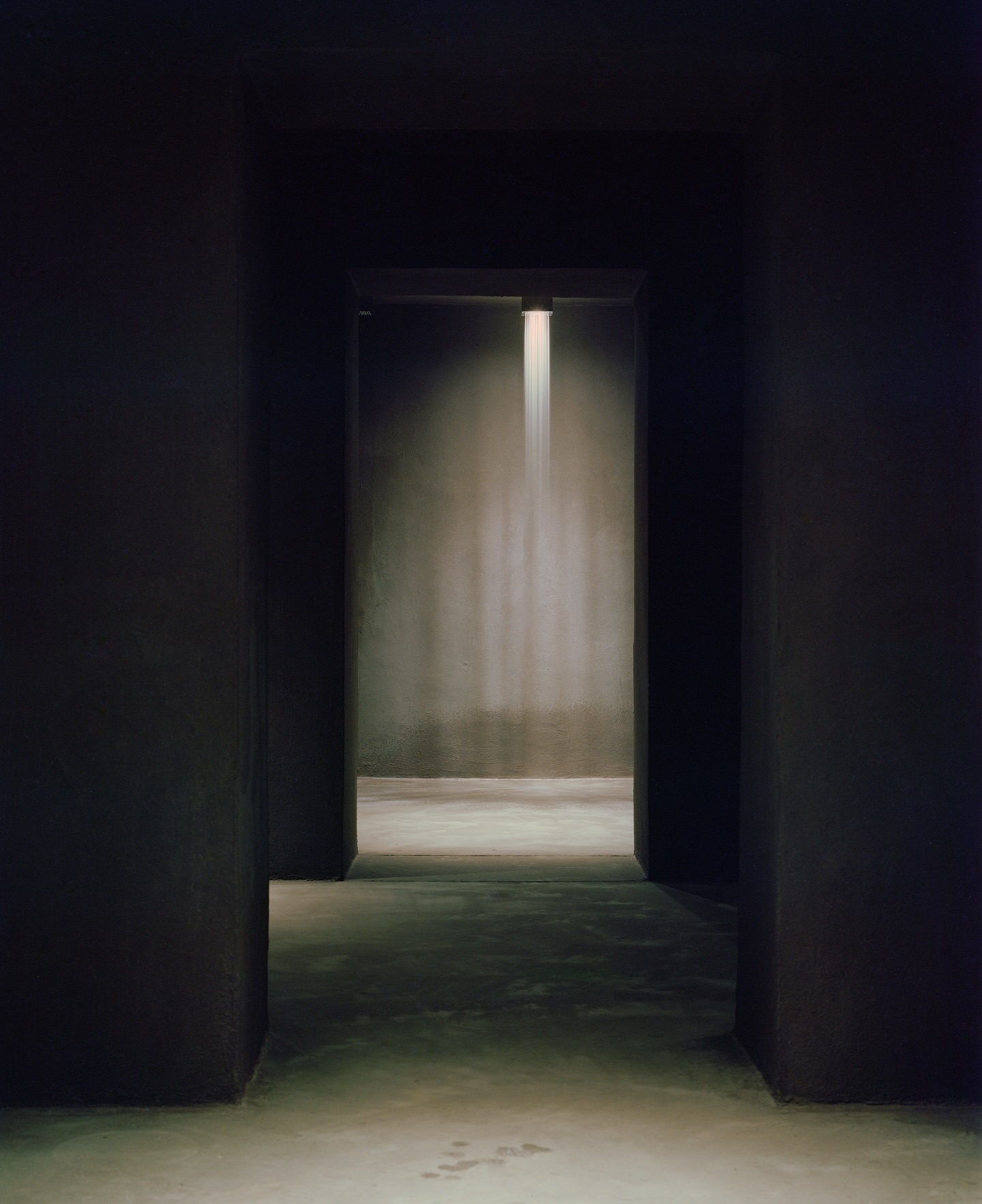 Riela by Oficina Satélite, photographed by Gregori Civera — Massive dark load-bearing walls open in line toward a lit chamber where a fine vertical curtain of water falls as vapor onto polished concrete.