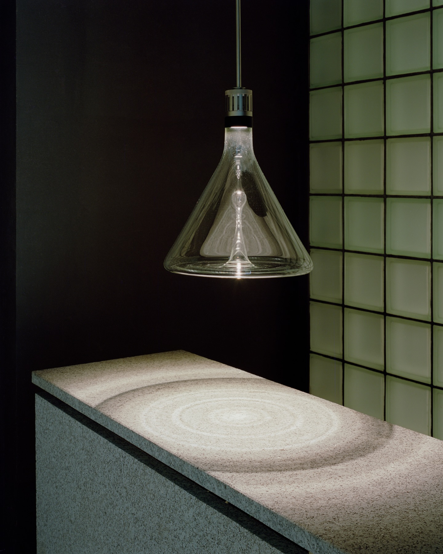 Riela by Oficina Satélite, photographed by Gregori Civera — A single hand-blown glass pendant hangs over a granite counter cut with concentric rings, the still trace of a dropped stone, against a pale-green glass-block wall.