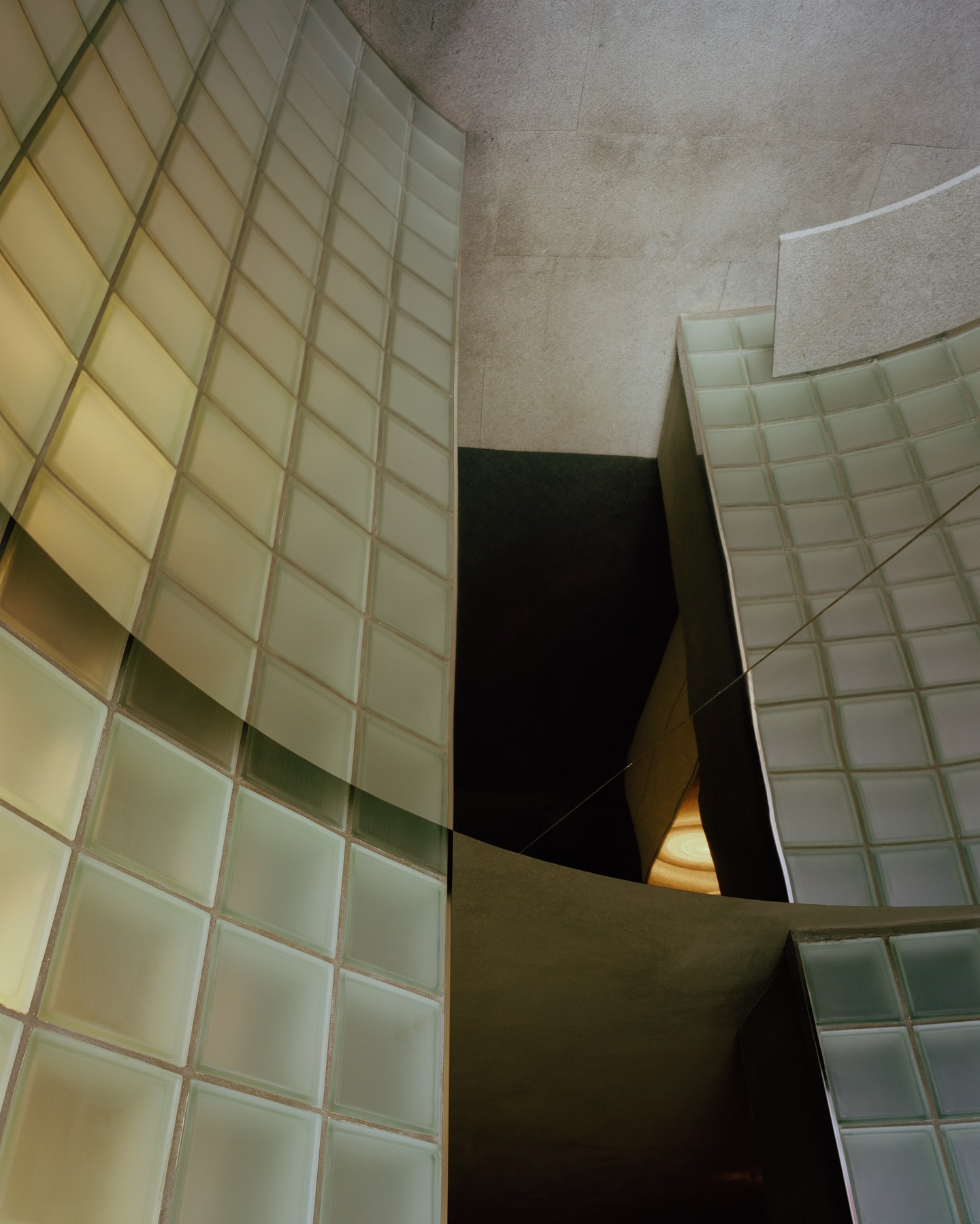 Riela by Oficina Satélite, photographed by Gregori Civera — Looking up into the stair void, curved backlit glass-block walls in pale green splay toward a raw concrete ceiling with a wedge of shadow opening between them.