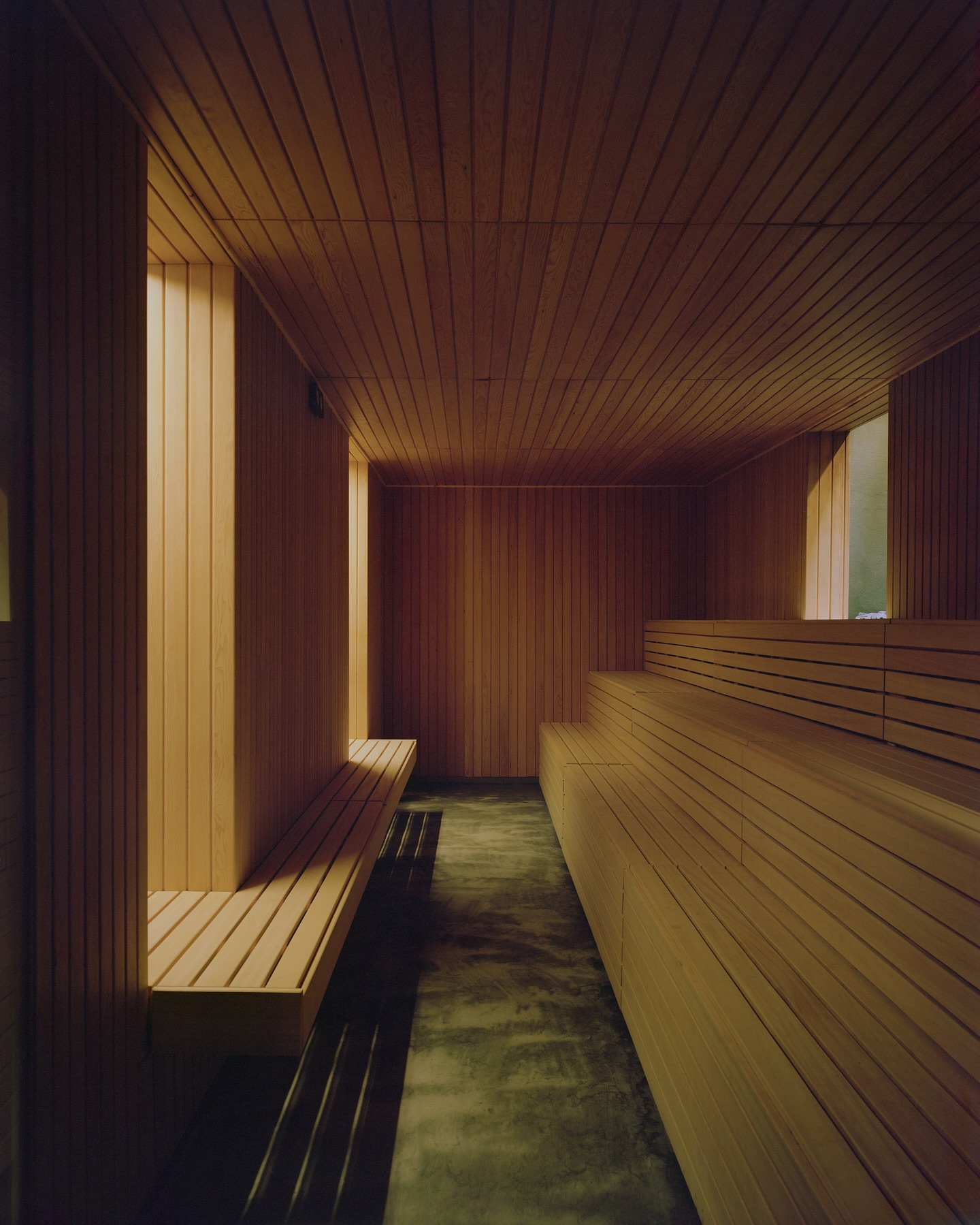 Riela by Oficina Satélite, photographed by Gregori Civera — The sauna lined entirely in pale slatted timber, tiered benches on one side and a floating bench on the other, a dark polished floor between them under warm concealed light.
