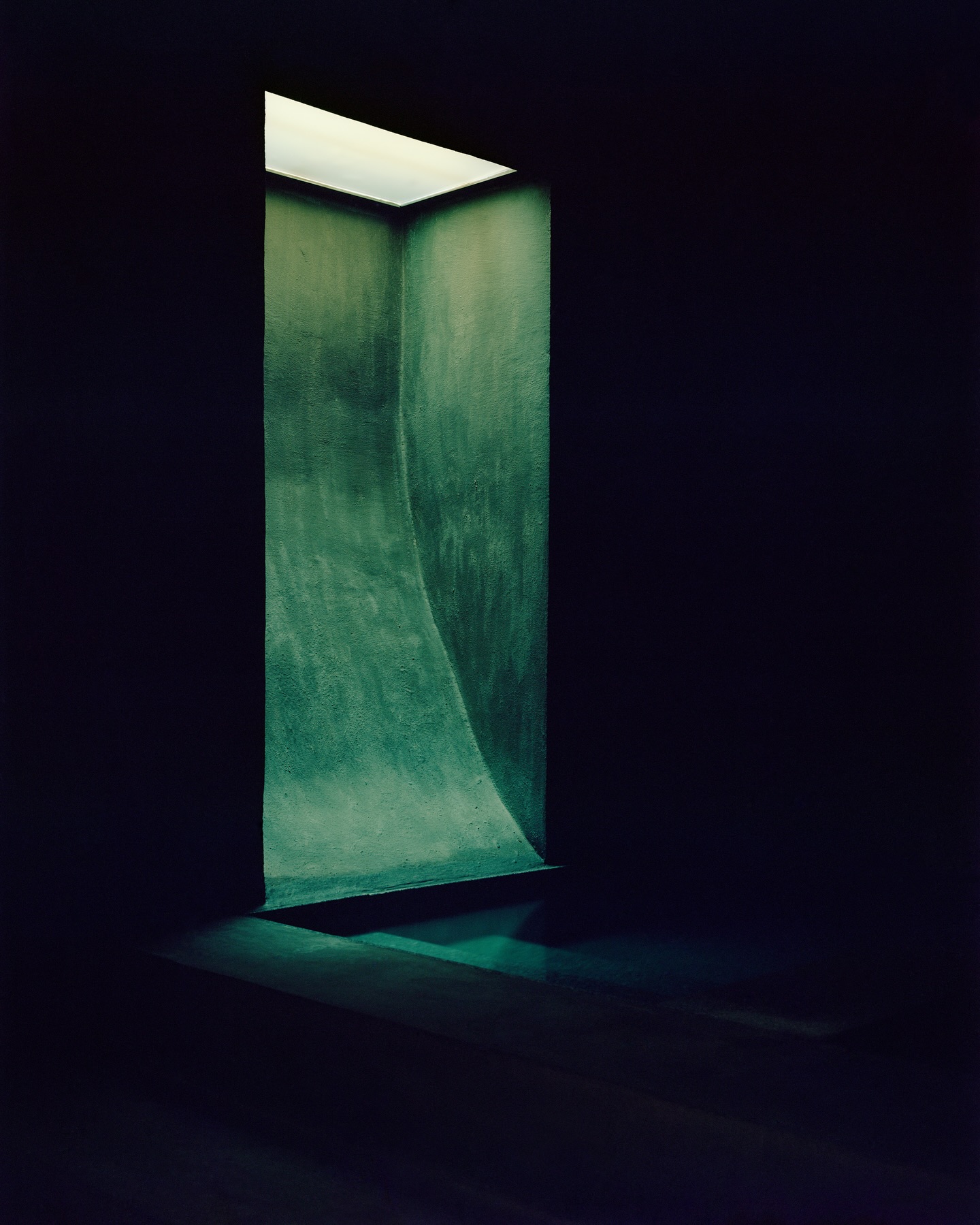 Riela by Oficina Satélite, photographed by Gregori Civera — A skylight drops a shaft of daylight down a curved dark-green plastered well, the pigment catching the light vertically against near-black surroundings.
