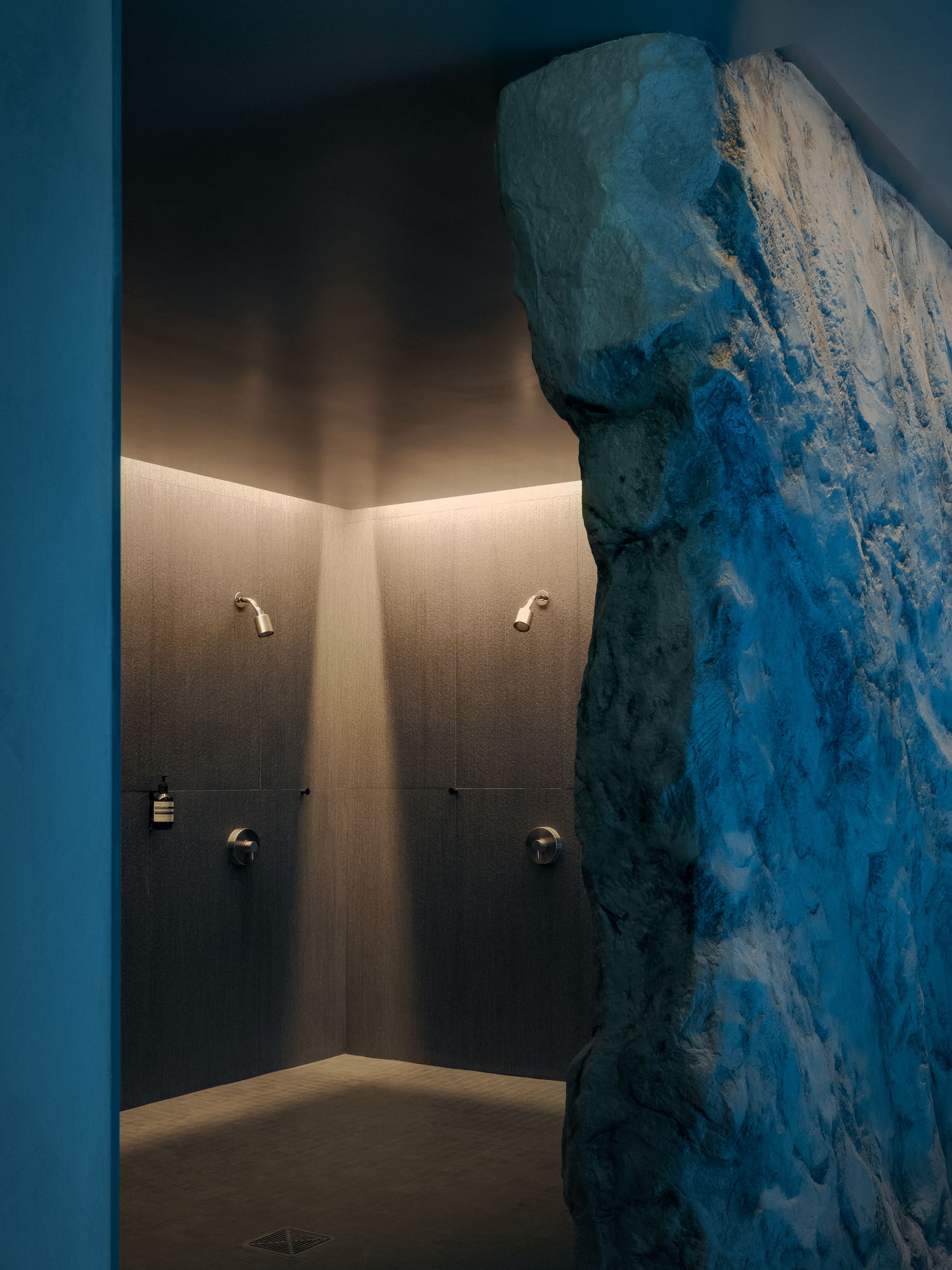 Recess by Future Simple Studio, photographed by Félix Michaud, Recess — Two nickel showerheads sit on dark textured tile, half-screened by a boulder-like wall lit an icy blue at the mouth of the shower room.