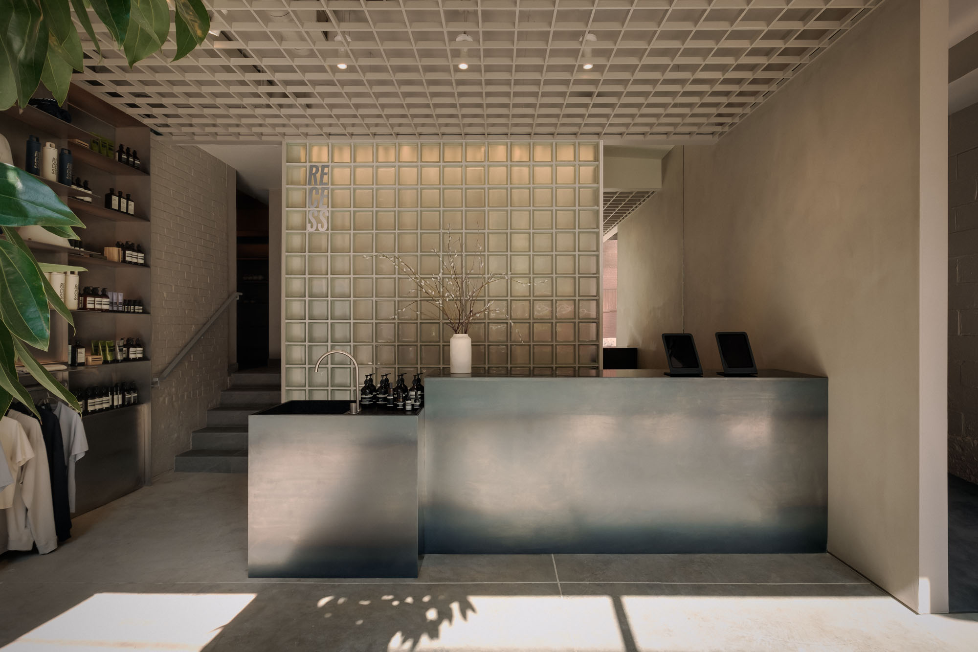 Recess by Future Simple Studio, photographed by Félix Michaud, Recess — A cold-rolled steel reception counter and lower hand basin stand before a glass-block lattice wall spelling RECESS, under an aluminium ceiling grid on polished concrete.