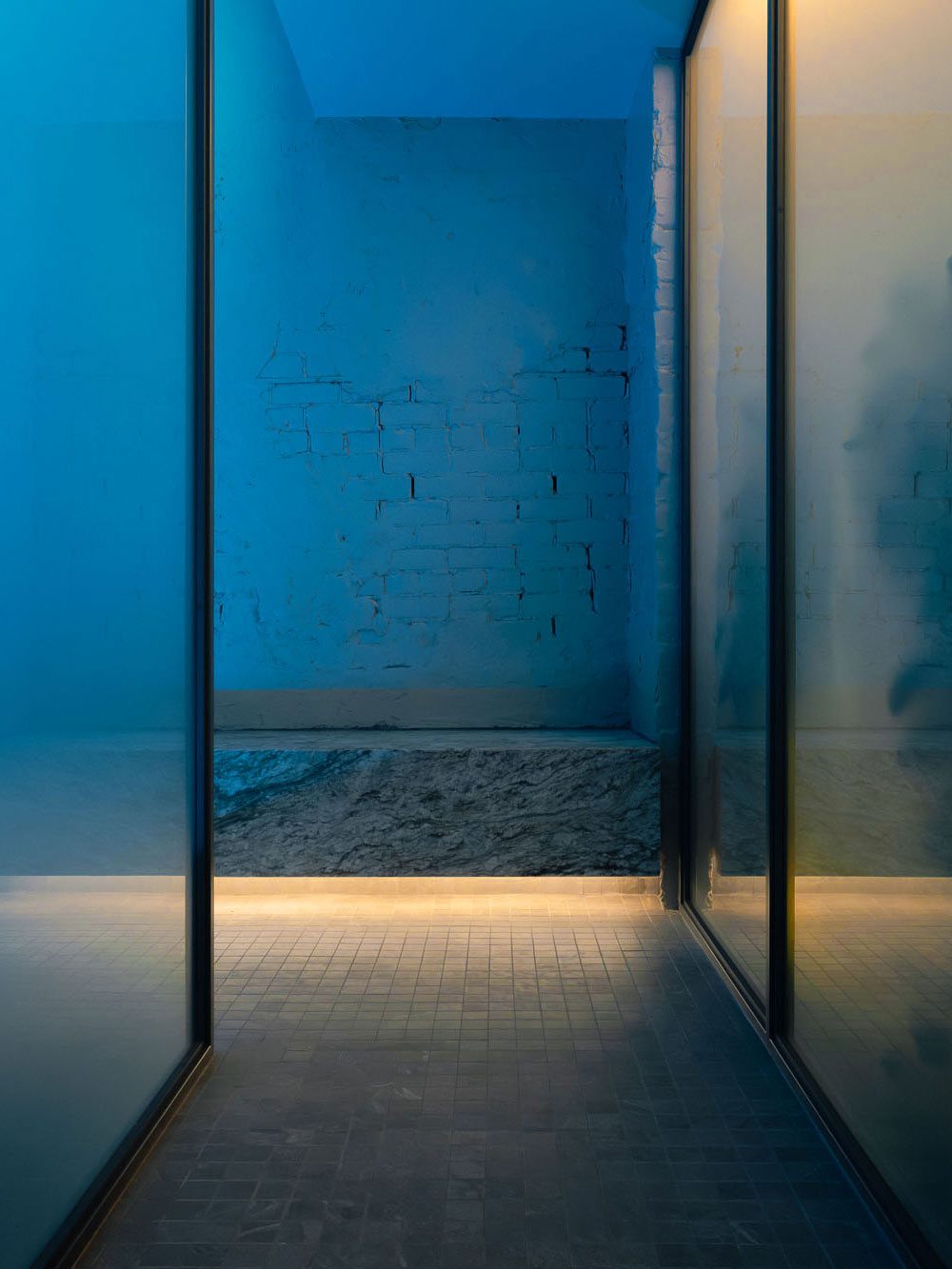 Recess by Future Simple Studio, photographed by Félix Michaud, Recess — Through frosted glass panels, a raw marble bench and cracked painted-brick wall glow deep blue above a grey mosaic floor.