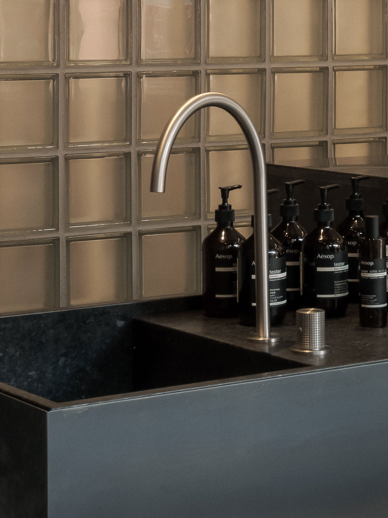 Recess by Future Simple Studio, photographed by Félix Michaud, Recess — A brushed steel gooseneck tap rises over a dark granite basin, a row of amber Aesop tester bottles lined up beside it against a warm glass-block wall.