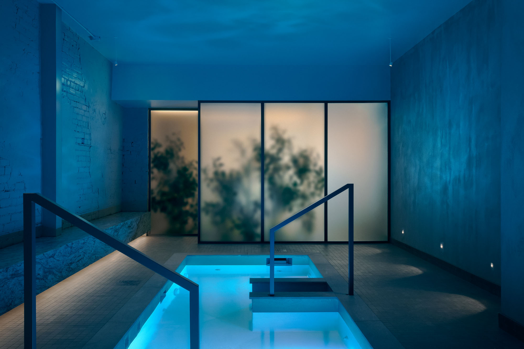 Recess by Future Simple Studio, photographed by Félix Michaud, Recess — Twin cold plunge pools glow glacial cyan in a blue room of painted brick and frosted glass, steel handrails leading down into the water.