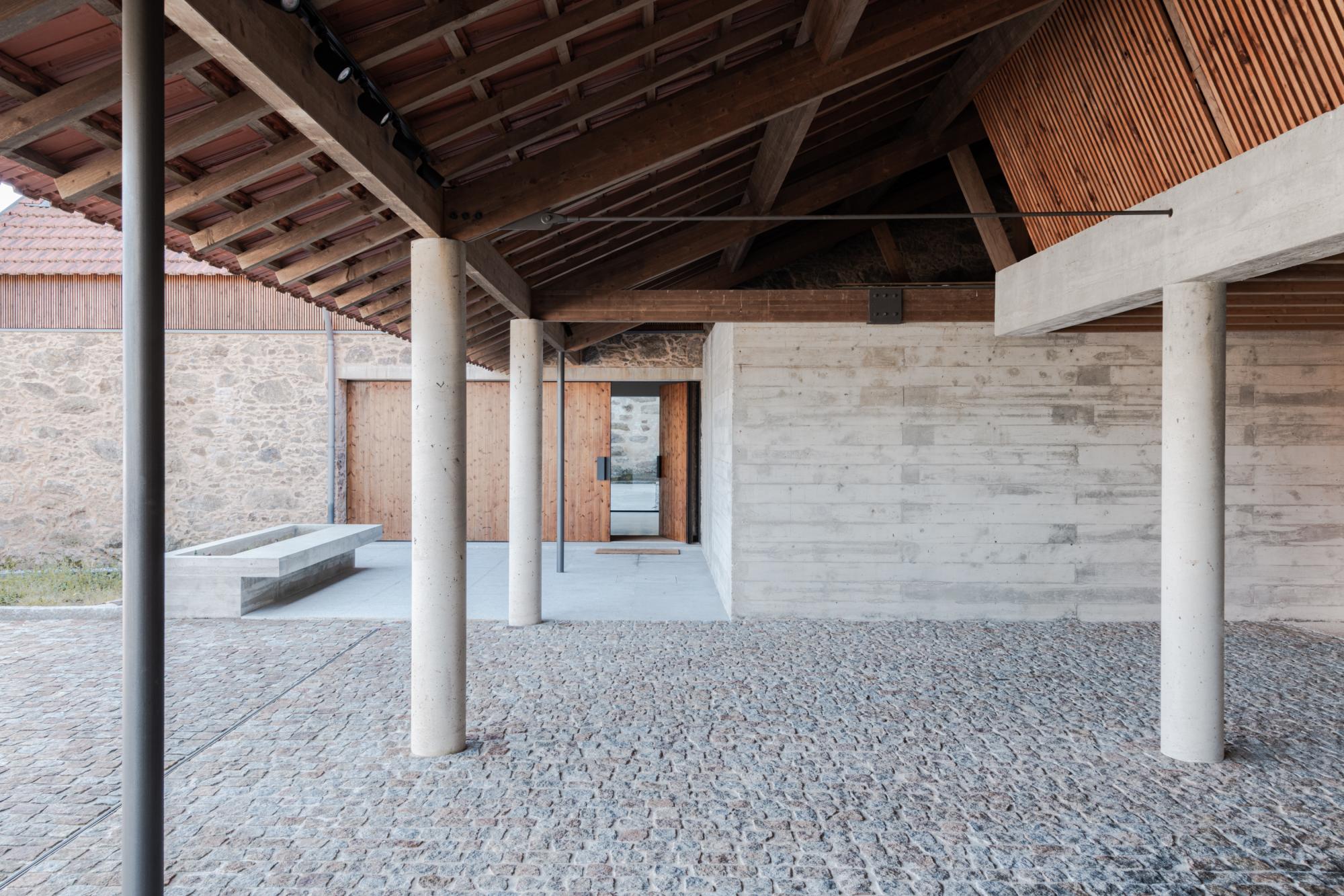 Single-Family Home by Branco del Rio Arquitectos, photographed by Frederico Martinho — A covered courtyard opens under exposed timber rafters carried on board-formed concrete columns, its granite sett floor meeting a horizontally shuttered concrete wall…
