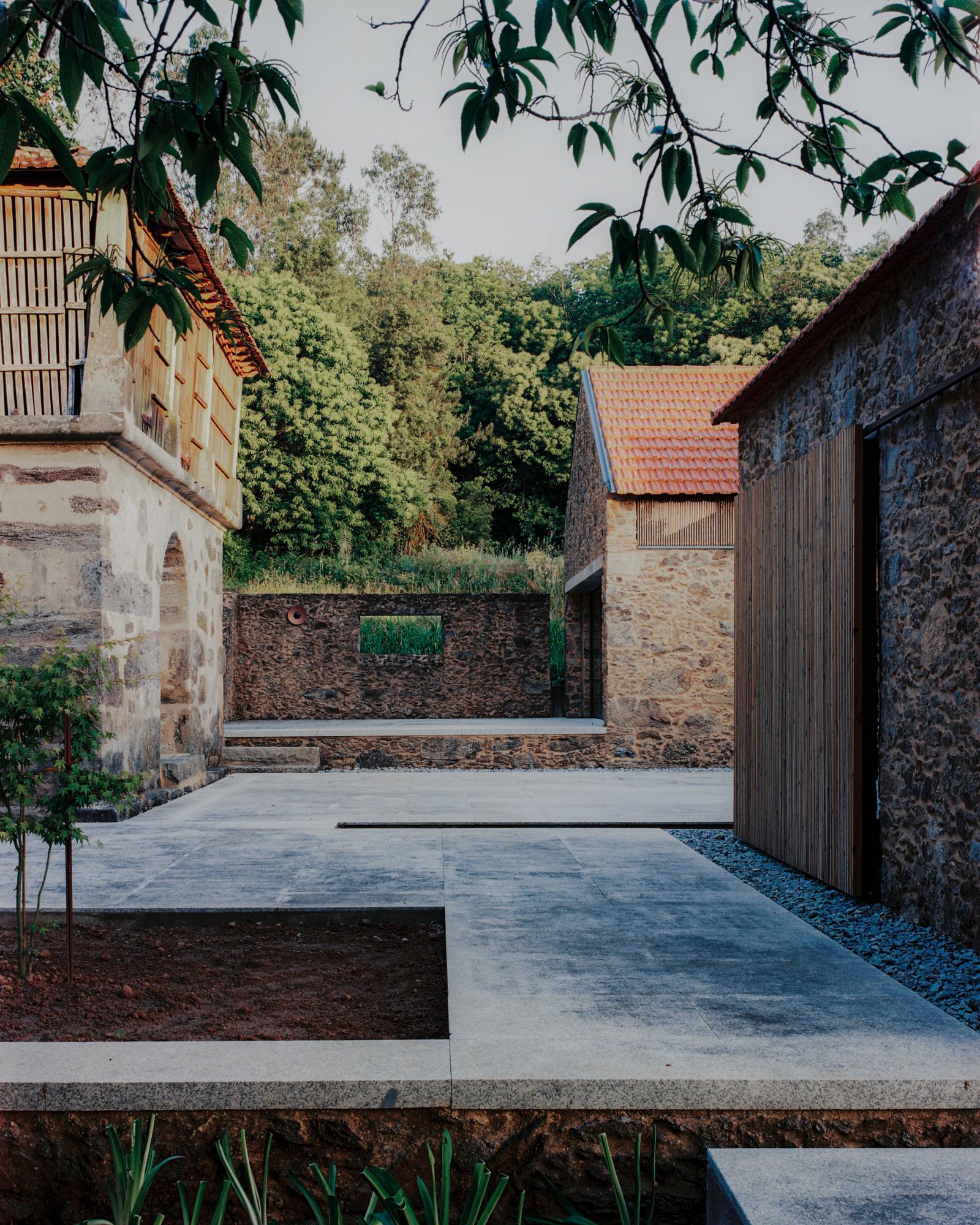 Single-Family Home by Branco del Rio Arquitectos, photographed by Frederico Martinho — A retained granite boulder sits in the foreground where a wide sliding glass wall opens the living room, pale sofas and a black kitchen block visible beneath a…