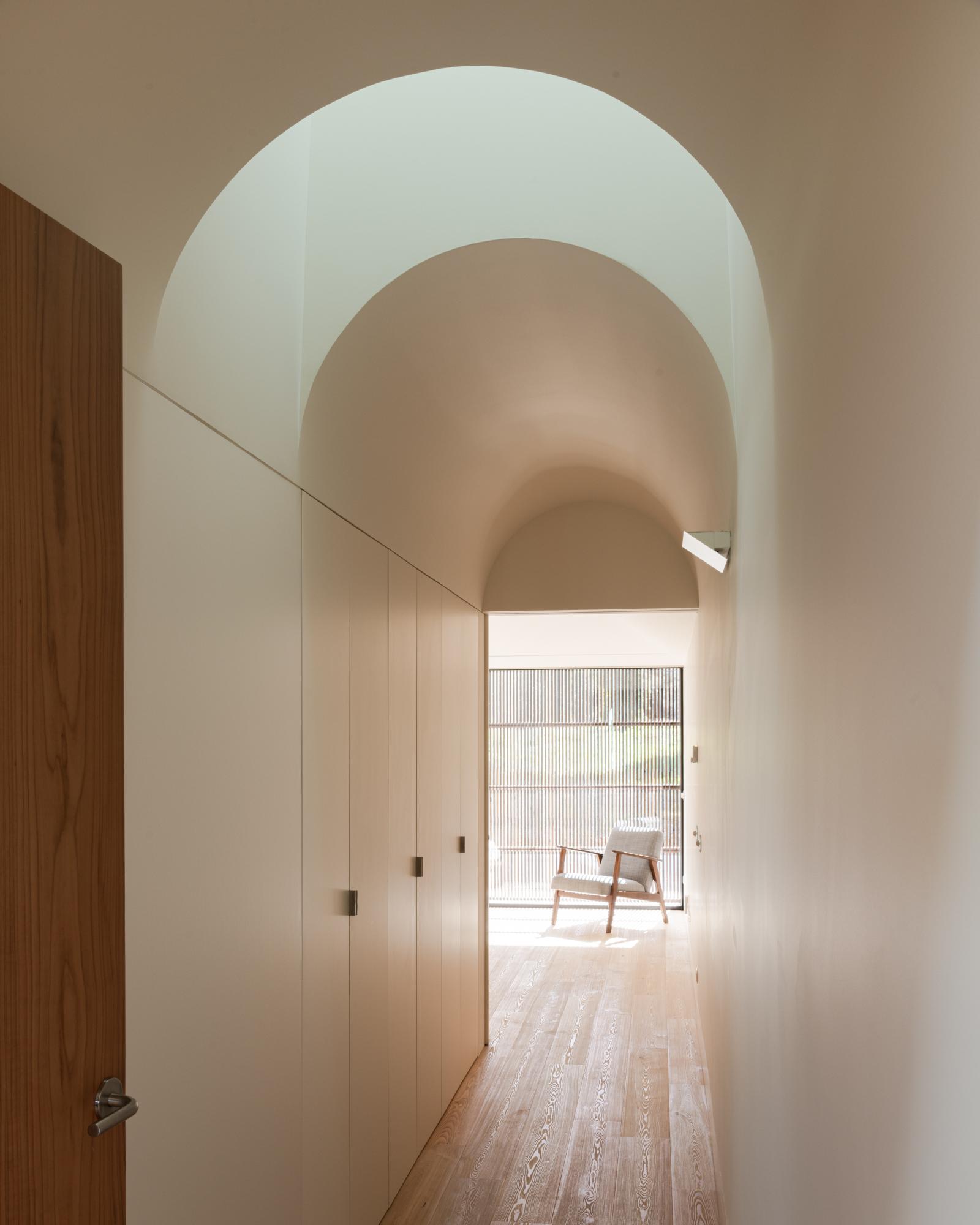 Single-Family Home by Branco del Rio Arquitectos, photographed by Frederico Martinho — Daylight pours down the white barrel vault of the bedroom-wing corridor, washing the built-in cabinetry and timber floor as a mid-century armchair waits by the…