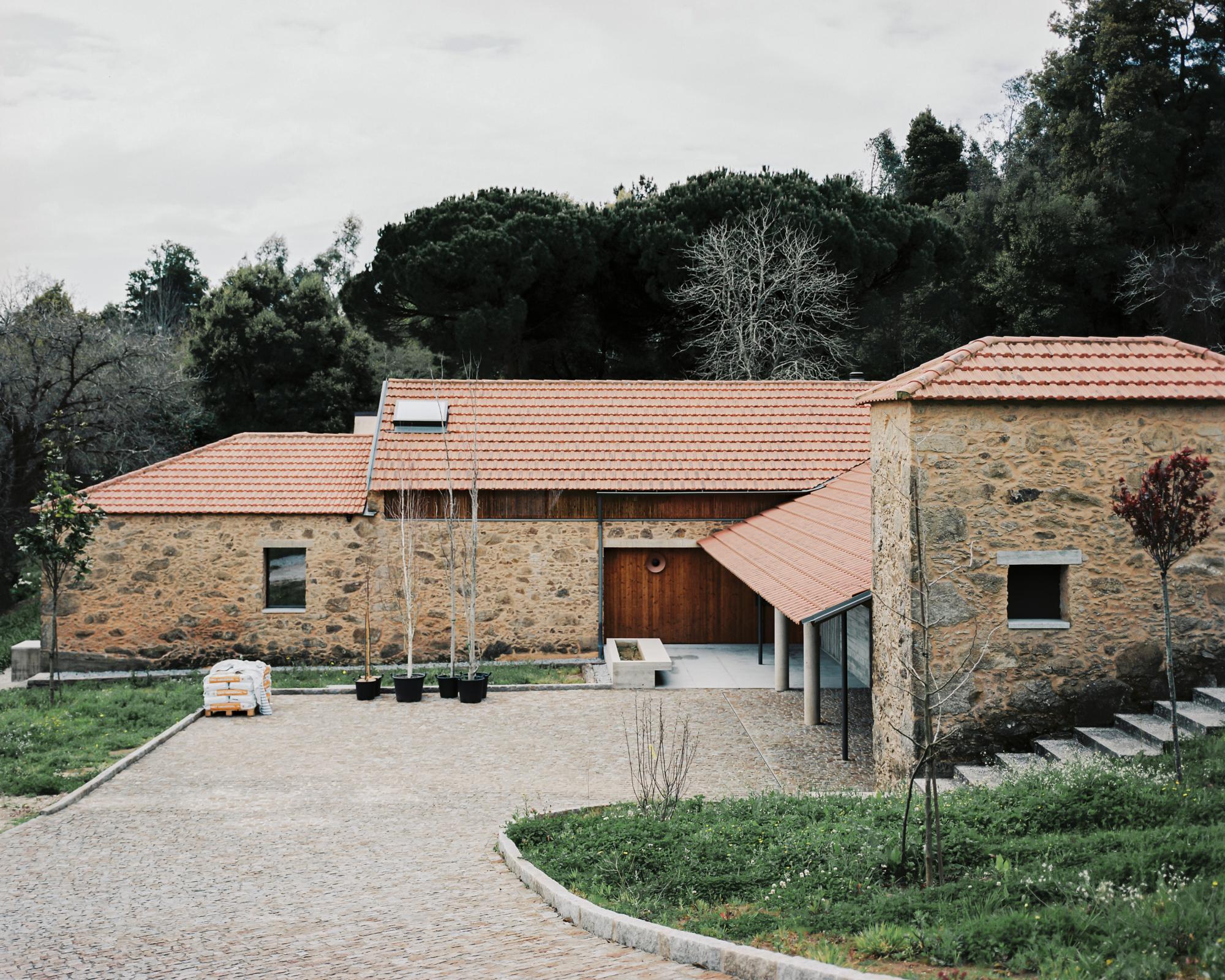 Single-Family Home by Branco del Rio Arquitectos, photographed by Frederico Martinho — The farmstead's front elevation stands in rough granite under terracotta tiles, a deep timber entrance door and a small pyramid-roofed volume set behind a cobblestone…