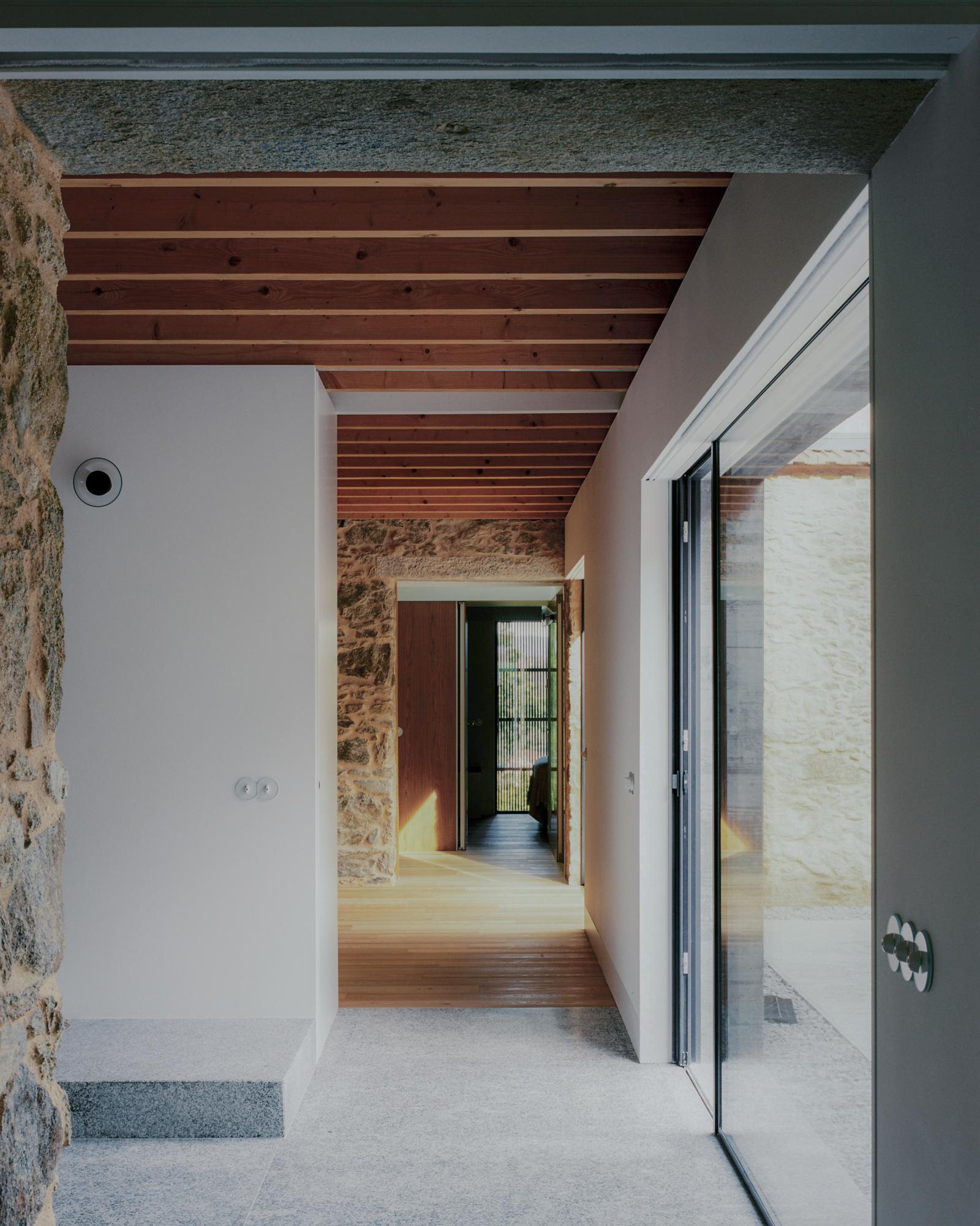Single-Family Home by Branco del Rio Arquitectos, photographed by Frederico Martinho — The dining room sits under a pitched timber joist ceiling, a black steel hearth and flue rising beside a walnut table, leather chairs and two white pendant lamps on a…