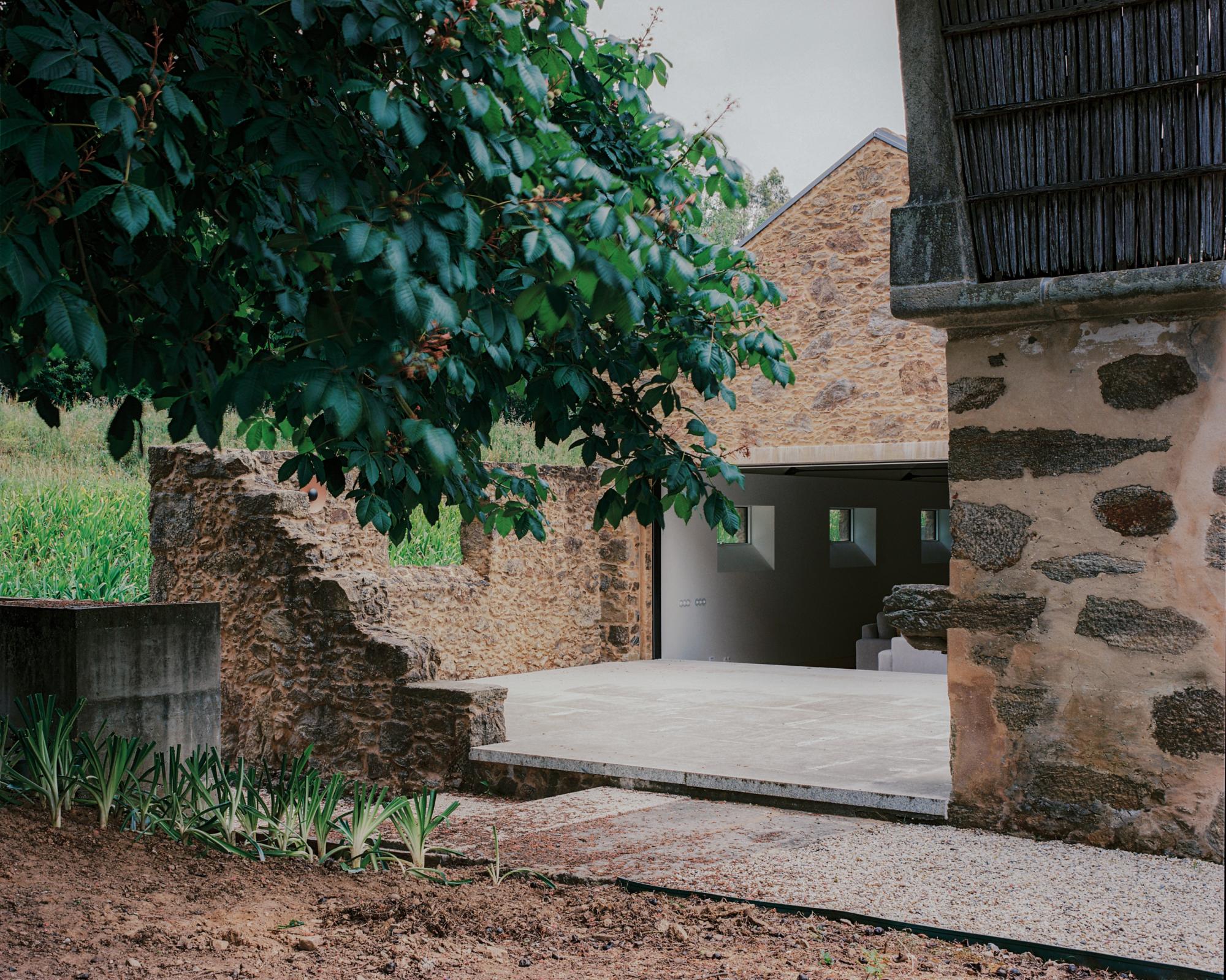 Single-Family Home by Branco del Rio Arquitectos, photographed by Frederico Martinho — A cherry tree shades a courtyard where rough granite walls, a low concrete bench and a poured platform frame a dark opening into the house, a ventilated timber eave…