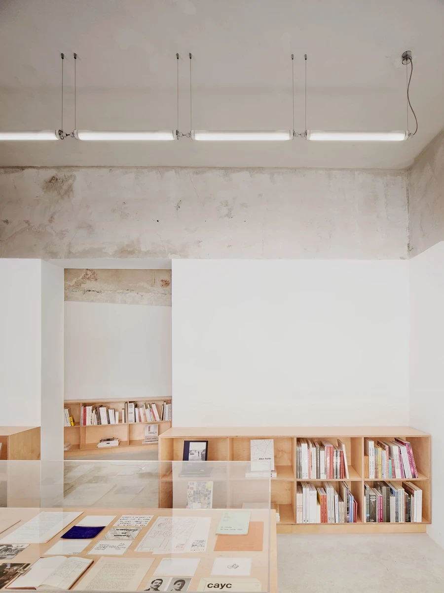 Lawrence Bookshop by Lluís Alexandre Casanovas Blanco, photographed by José Hevia — A broad blank white wall opens at left to a book-lined back room, a low birch shelf and the edge of an acrylic vitrine below the unrepaired plaster band.
