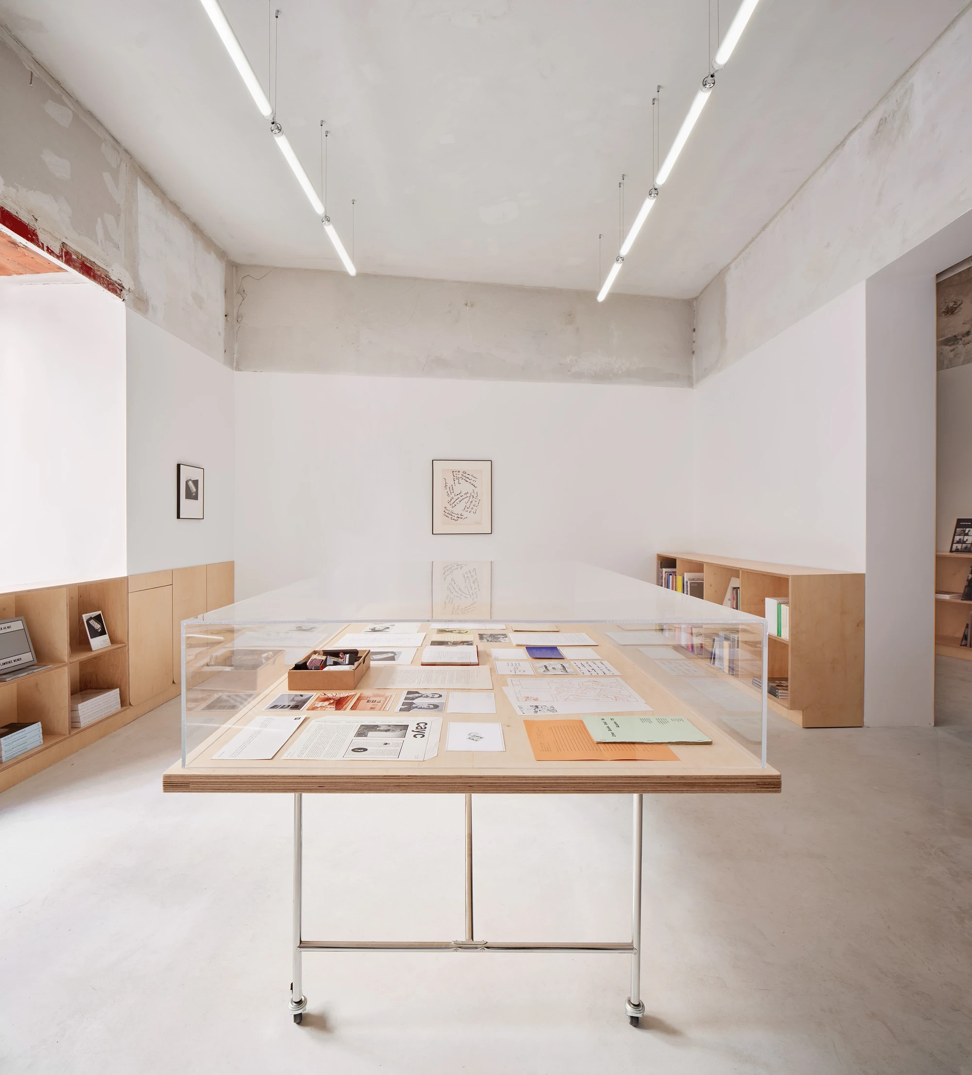 Lawrence Bookshop by Lluís Alexandre Casanovas Blanco, photographed by José Hevia — The long vitrine table fills the room under suspended fluorescent tubes, a single framed drawing centred on the white wall above the raw plaster line.