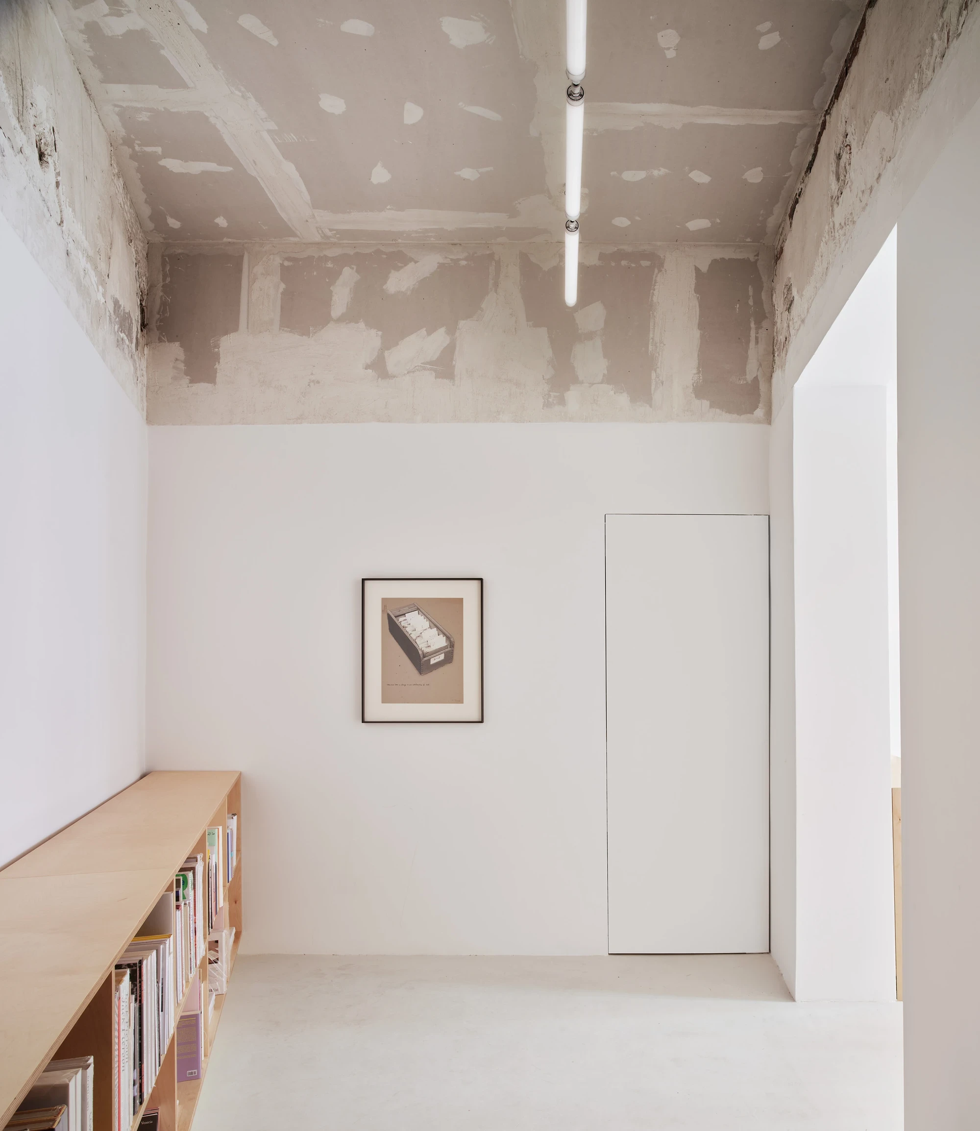 Lawrence Bookshop by Lluís Alexandre Casanovas Blanco, photographed by José Hevia — The back room keeps its patched drywall and plaster ceiling, a framed print centred above a low birch bookshelf beside a flush plywood door.
