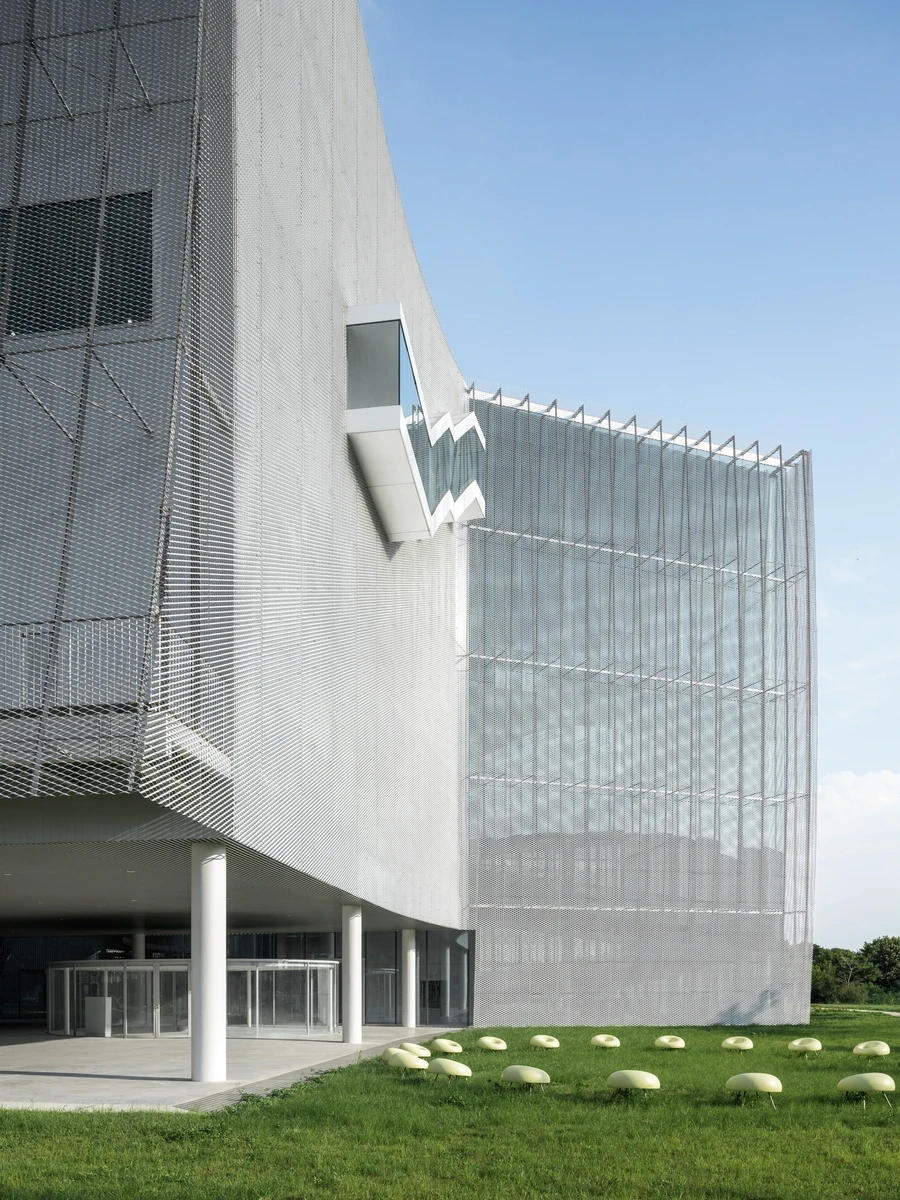 Taichung Art Museum by SANAA, photographed by Yi-Hsien Lee (YHLAA) — Silver expanded metal mesh and vertical glass fins wrap a faceted white volume lifted on slender columns, with pale egg-shaped stones scattered across the lawn below.