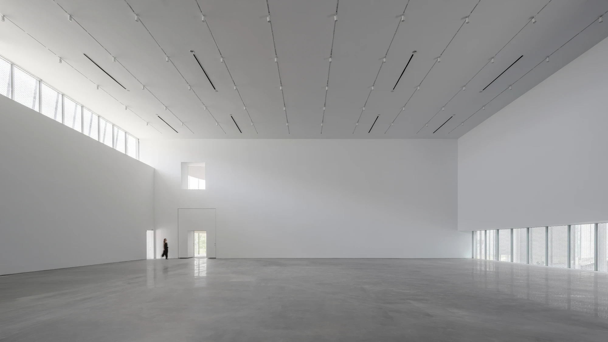 Taichung Art Museum by SANAA, photographed by Yi-Hsien Lee (YHLAA) — A double-height gallery holds tall white walls, track lighting and a polished concrete floor, lit by clerestory windows screened in metal mesh.