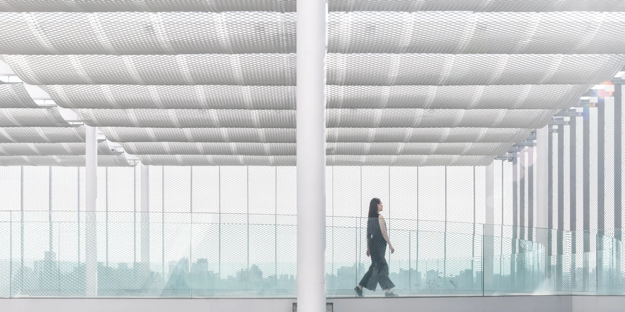 Taichung Art Museum by SANAA, photographed by Yi-Hsien Lee (YHLAA) — An elevated walkway passes under a white expanded-metal-mesh soffit, the hazy city skyline dissolving behind the screen.