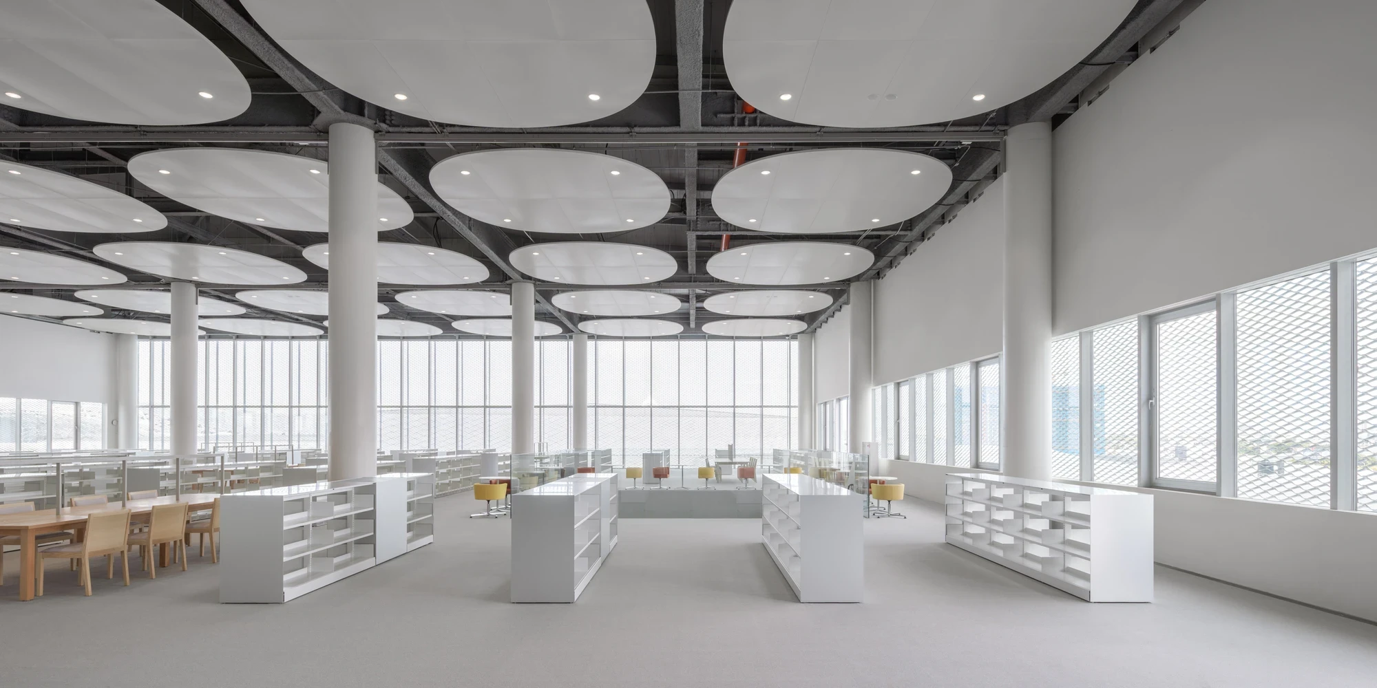 Taichung Art Museum by SANAA, photographed by Yi-Hsien Lee (YHLAA) — The library reading room spreads under floating oval acoustic clouds, with low white shelving, pale wood tables and yellow and red chairs beside mesh-screened windows.