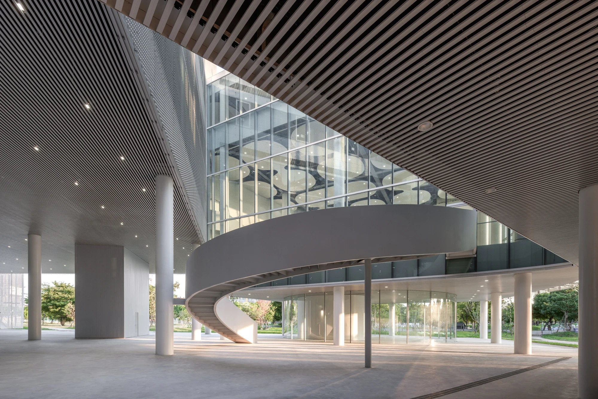 Taichung Art Museum by SANAA, photographed by Yi-Hsien Lee (YHLAA) — In the shaded undercroft, a black timber-slatted soffit angles over a white spiral ramp and slender columns opening to the park.