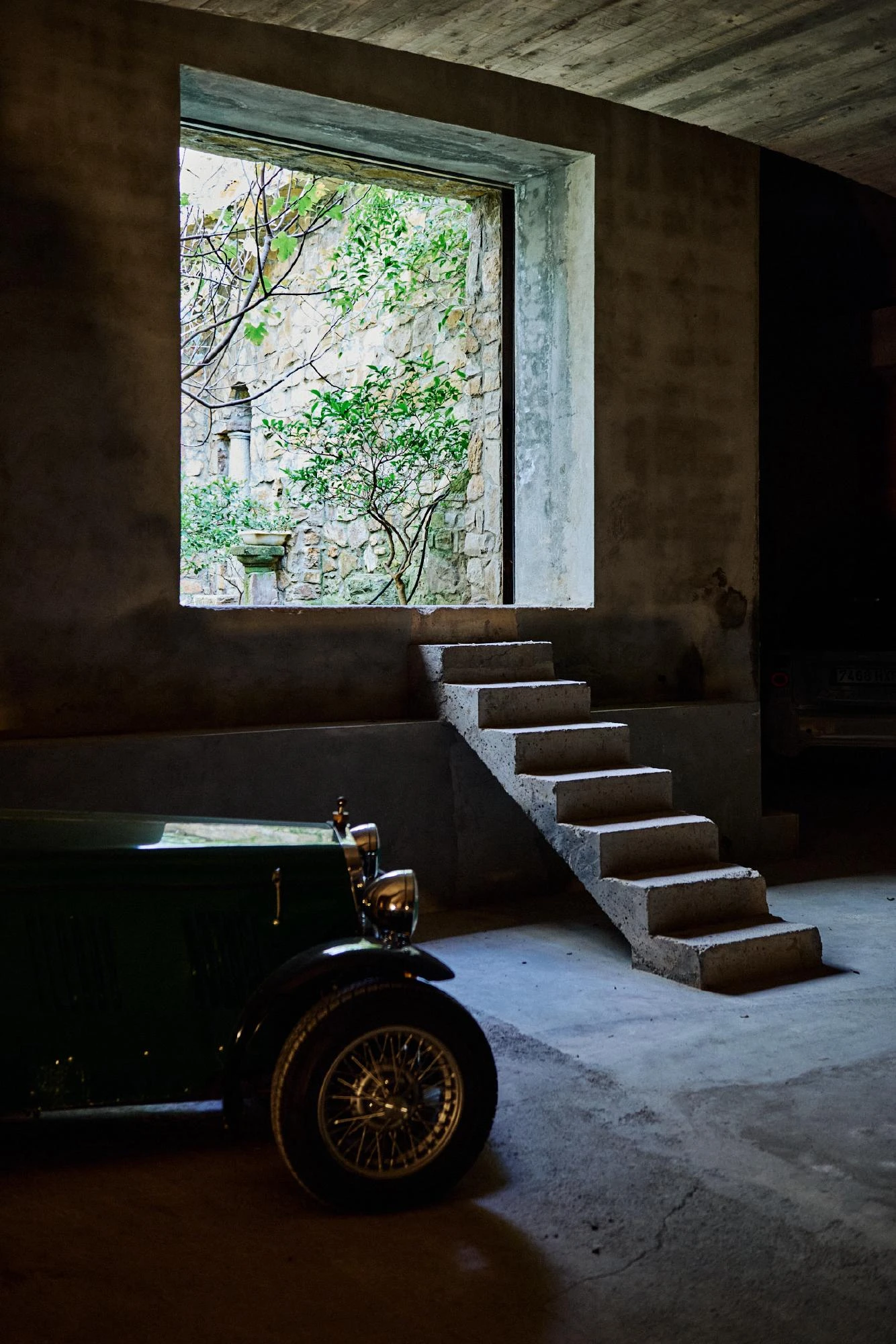 Xavier Corberó House and Studio by Xavier Corberó, photographed by Daniel Schäfer — A green vintage roadster with wire wheels sits in a dark garage beside a raw concrete stair, a square window cut to a wall of rough stone and greenery.