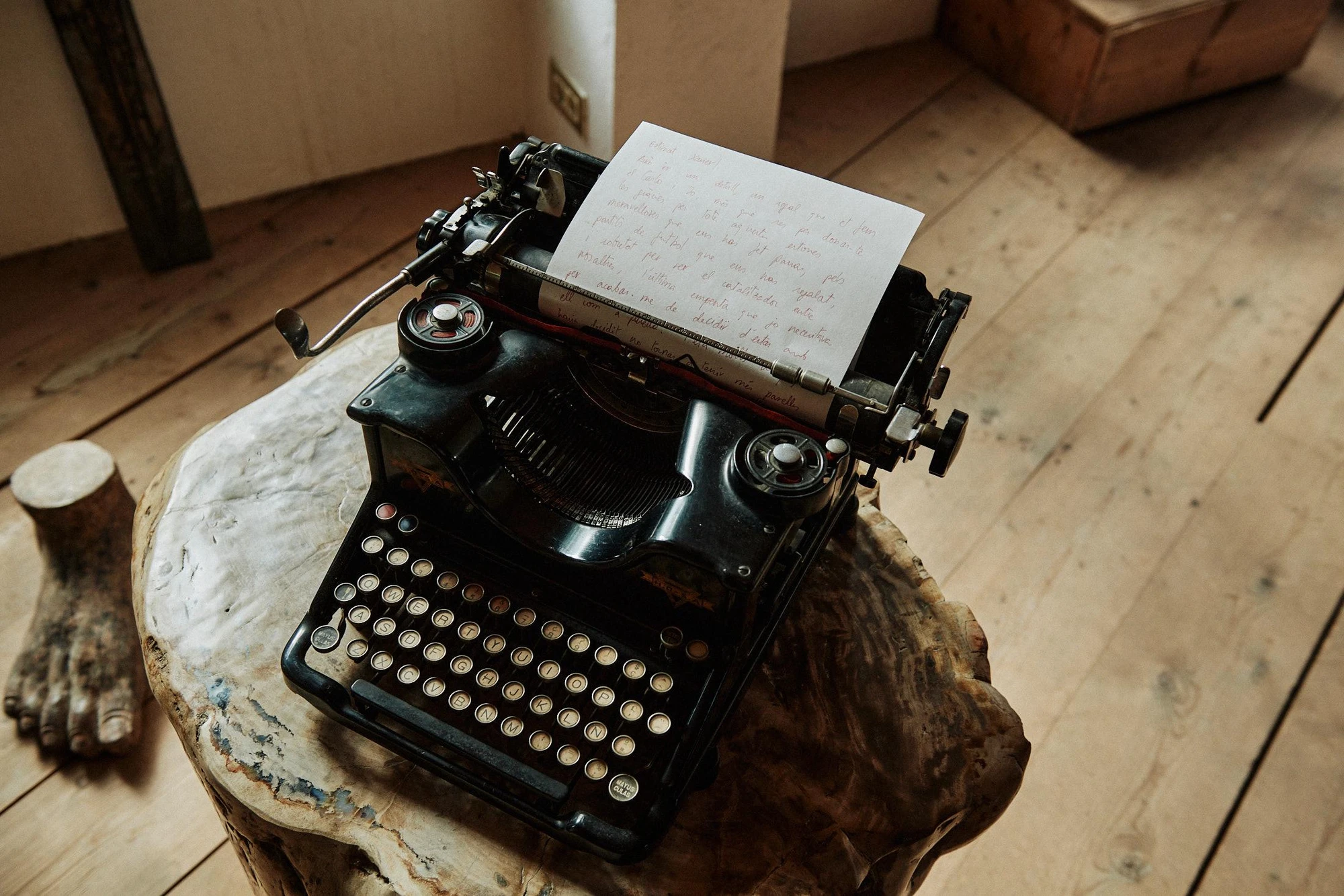 Xavier Corberó House and Studio by Xavier Corberó, photographed by Daniel Schäfer — A black vintage typewriter with a handwritten Catalan letter in its roller rests on a raw timber stump, a small carved stone foot on the plank floor beside it.