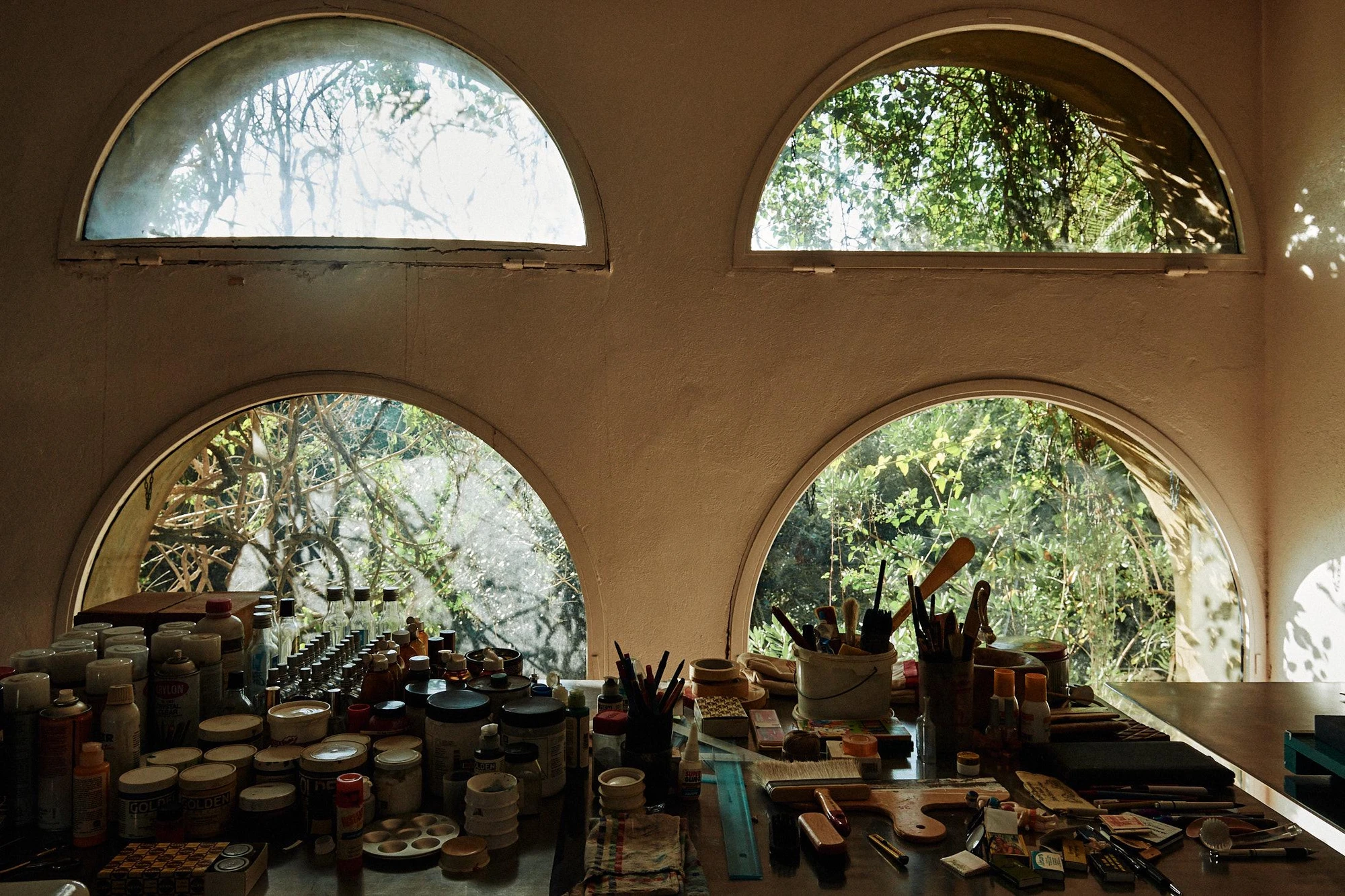 Xavier Corberó House and Studio by Xavier Corberó, photographed by Daniel Schäfer — A studio worktable crowded with jars of pigment and pots of brushes sits beneath two arched windows filled with tangled greenery.